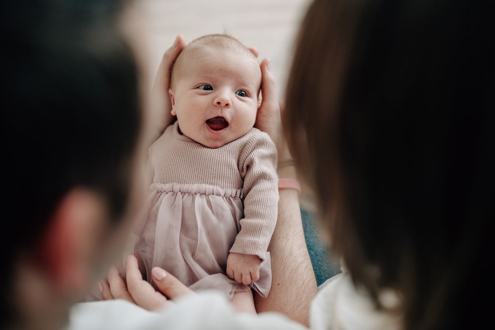 A baby wearing a light colored dress yawns while being held by someone.Newborn Fotografie Frankfurt