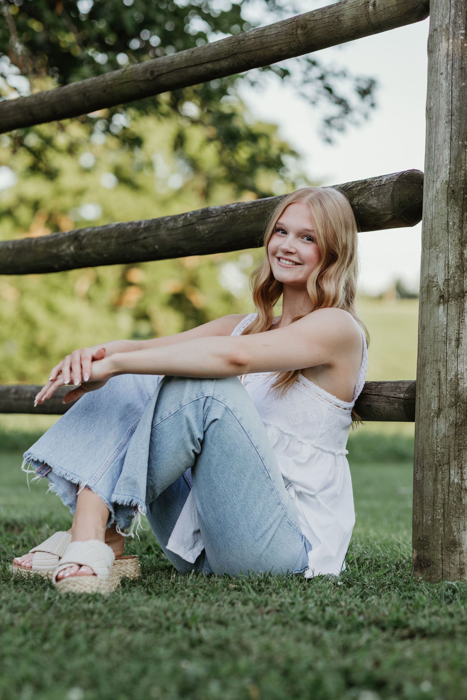 A senior wearing jeans and a white top sits casually against a rustic wooden fence in a grassy outdoor setting.