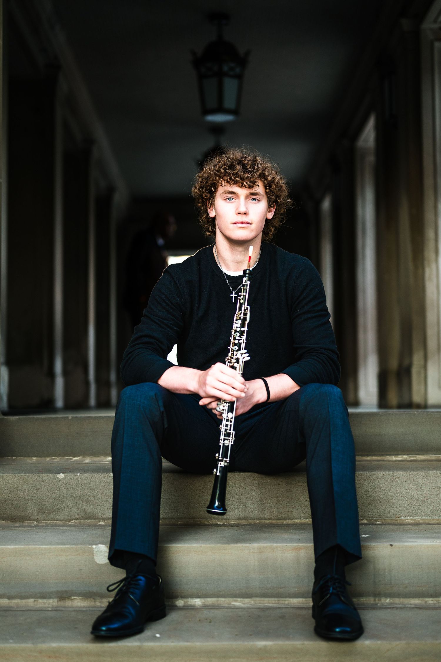 Musician sits on stone steps holding oboe in classical architecture setting.