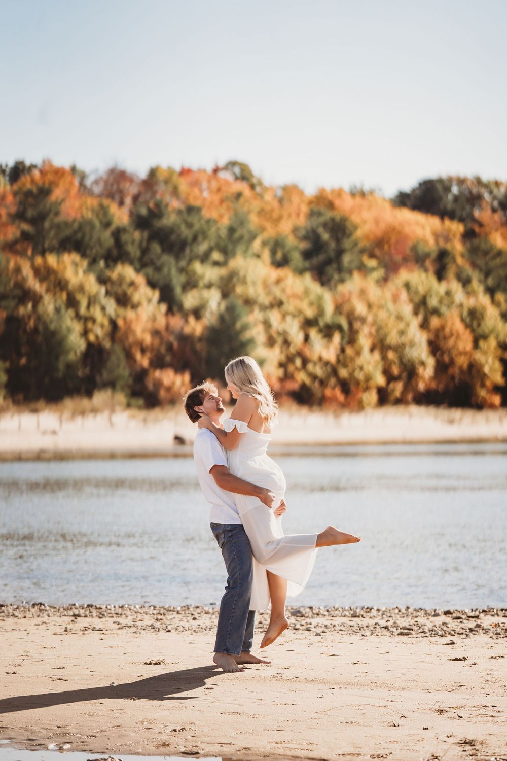 A couple embraces and spins on a sandy beach with autumn foliage in the background during golden hour.