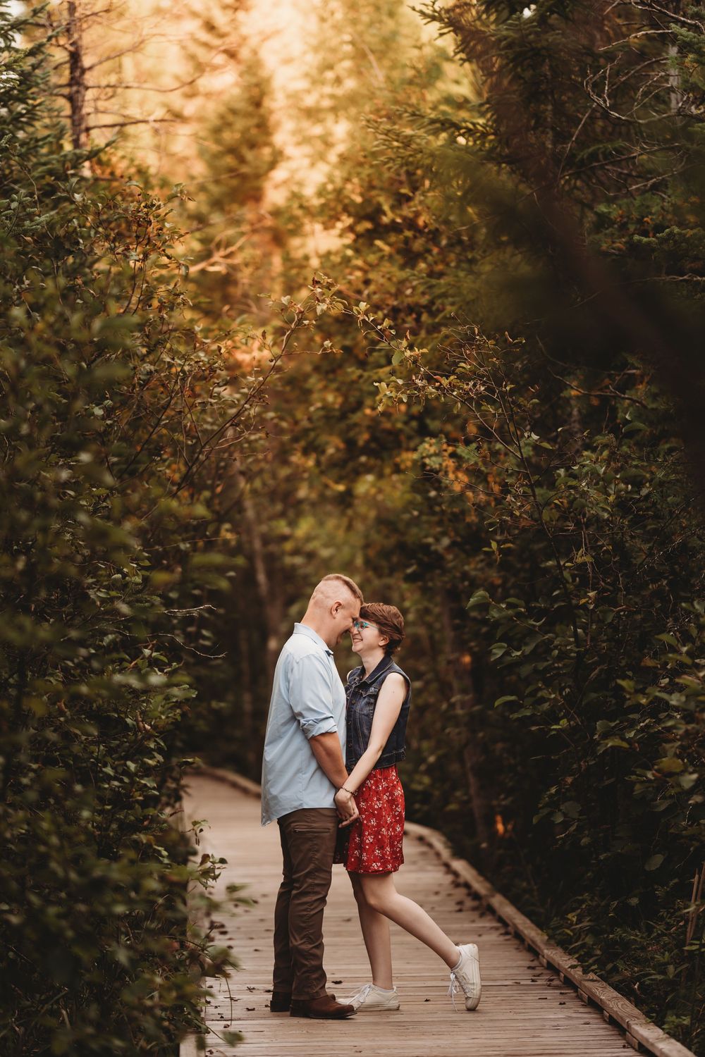 A romantic couple shares an intimate moment on a wooden bridge surrounded by lush greenery at sunset.