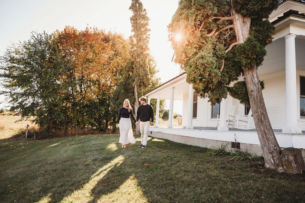 A white farmhouse with a wrap-around porch sits nestled among autumn trees at sunset.