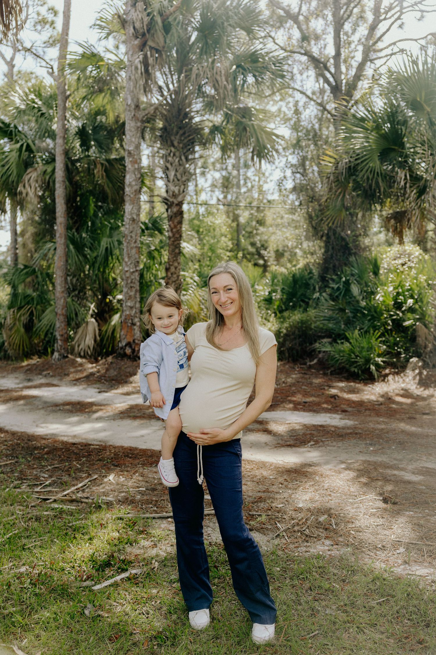 Pregnant woman in beige top and navy flares holds toddler boy outdoors among lush palm trees in a maternity photo.