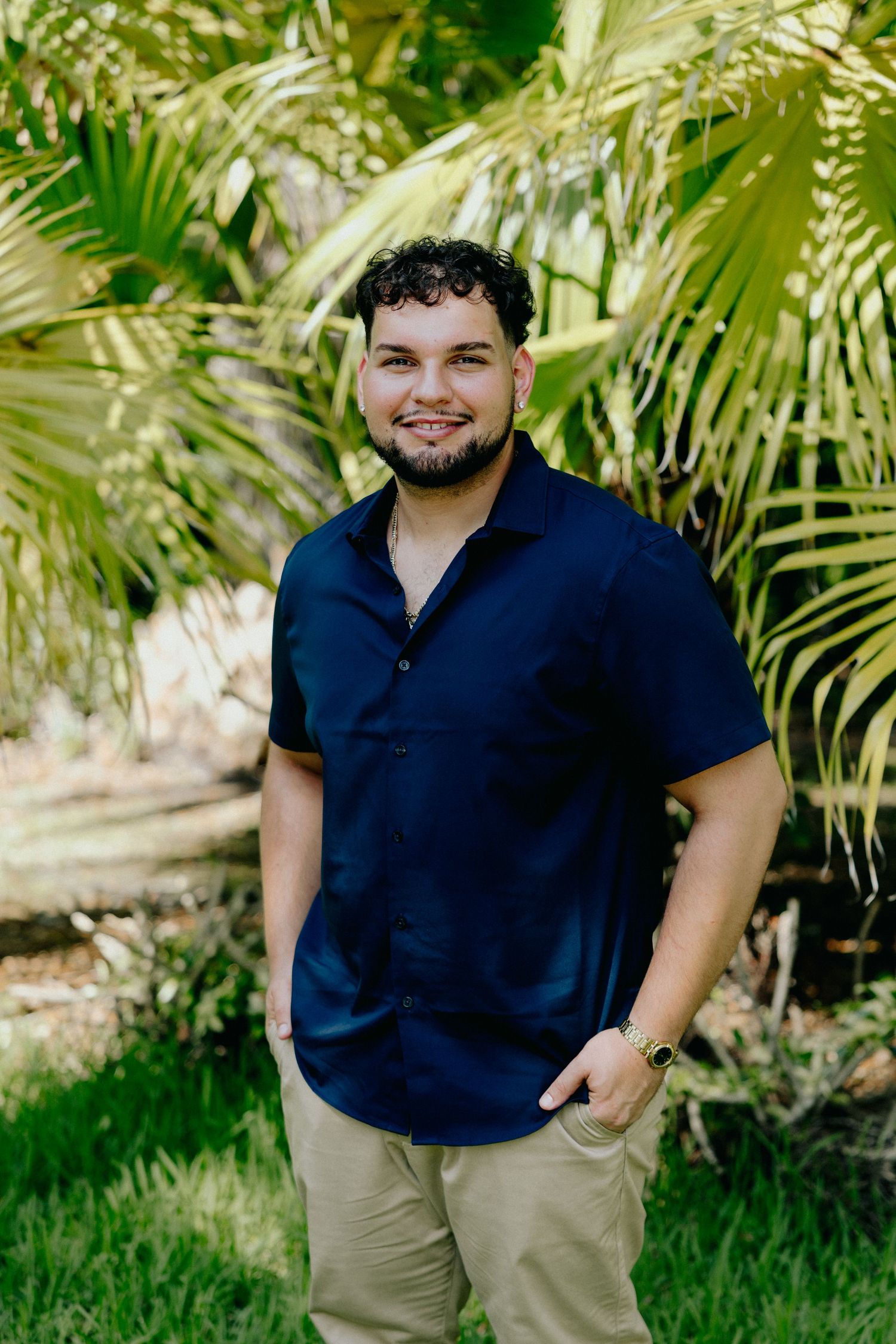 Smiling young man in navy polo shirt posing before tropical palm foliage.