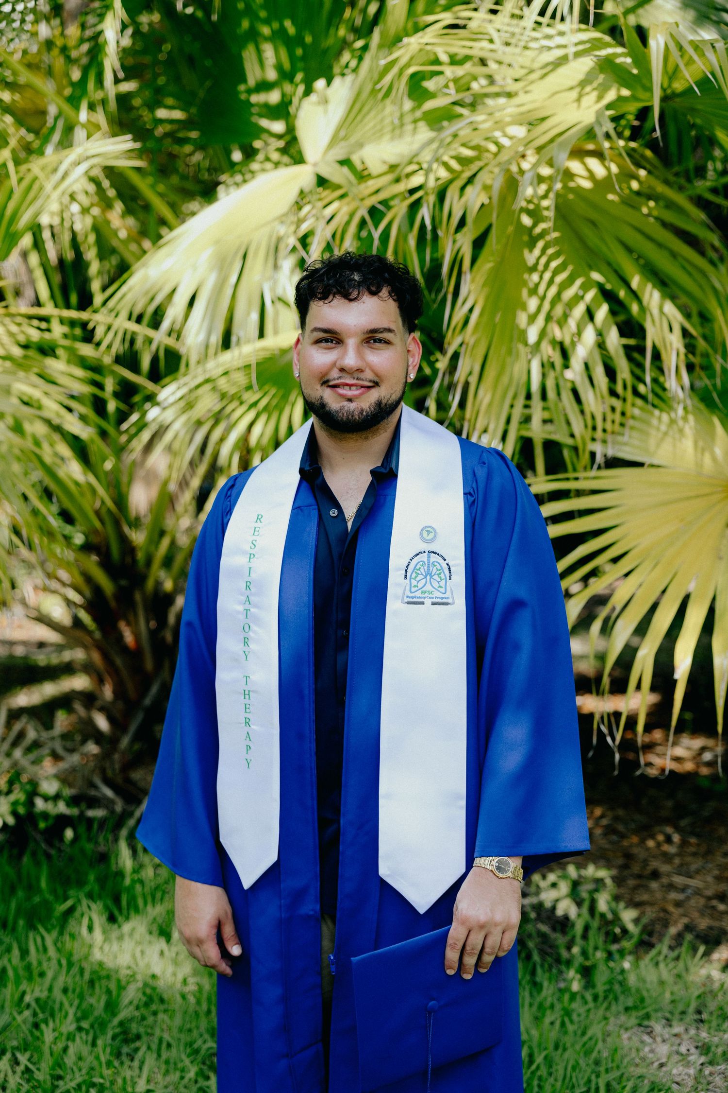 Smiling graduate in blue cap and gown with white stole before palm trees.