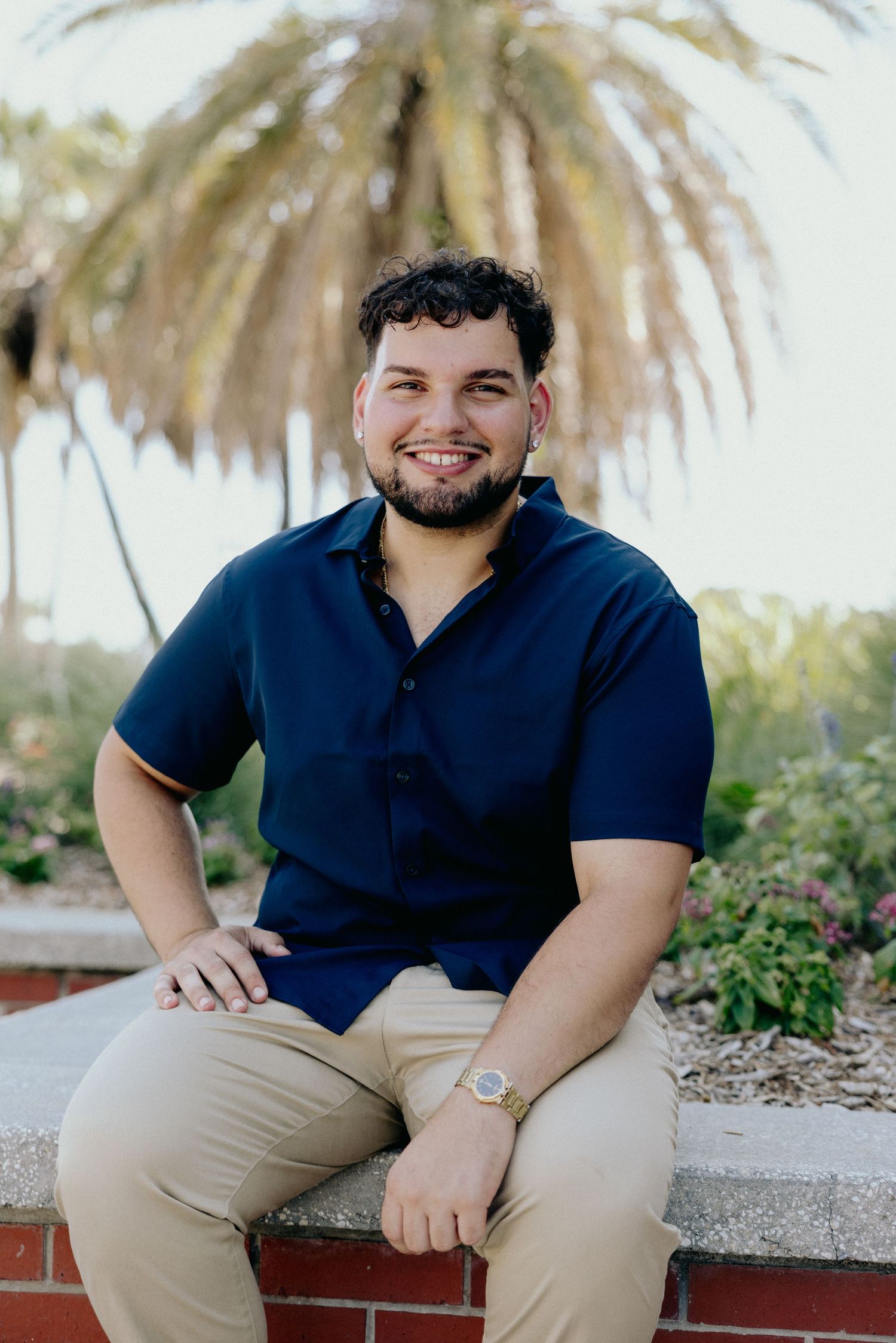 Smiling young man in navy shirt seated on brick wall with palm tree backdrop.