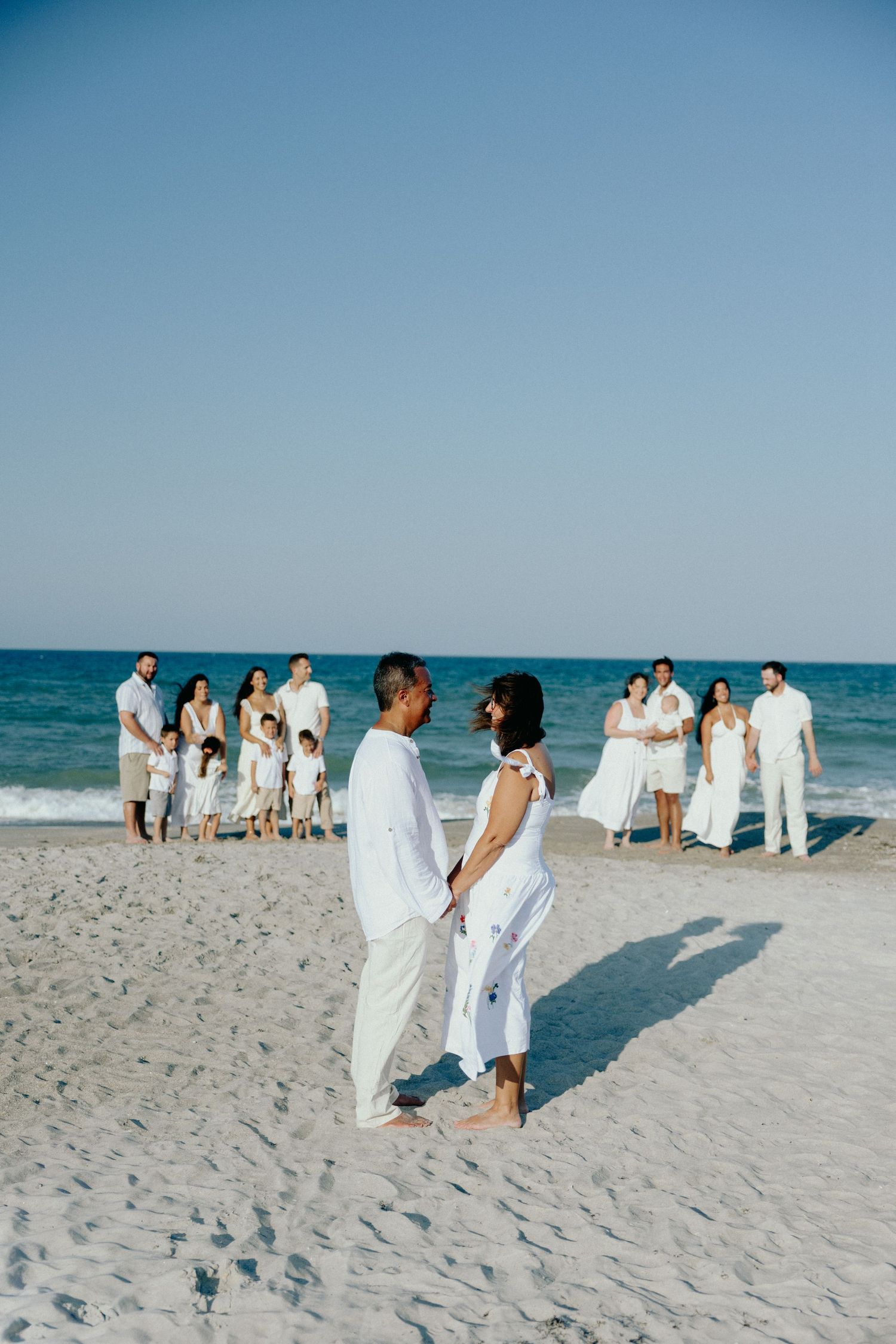 Family group in all-white outfits posing on sandy beach with ocean backdrop.