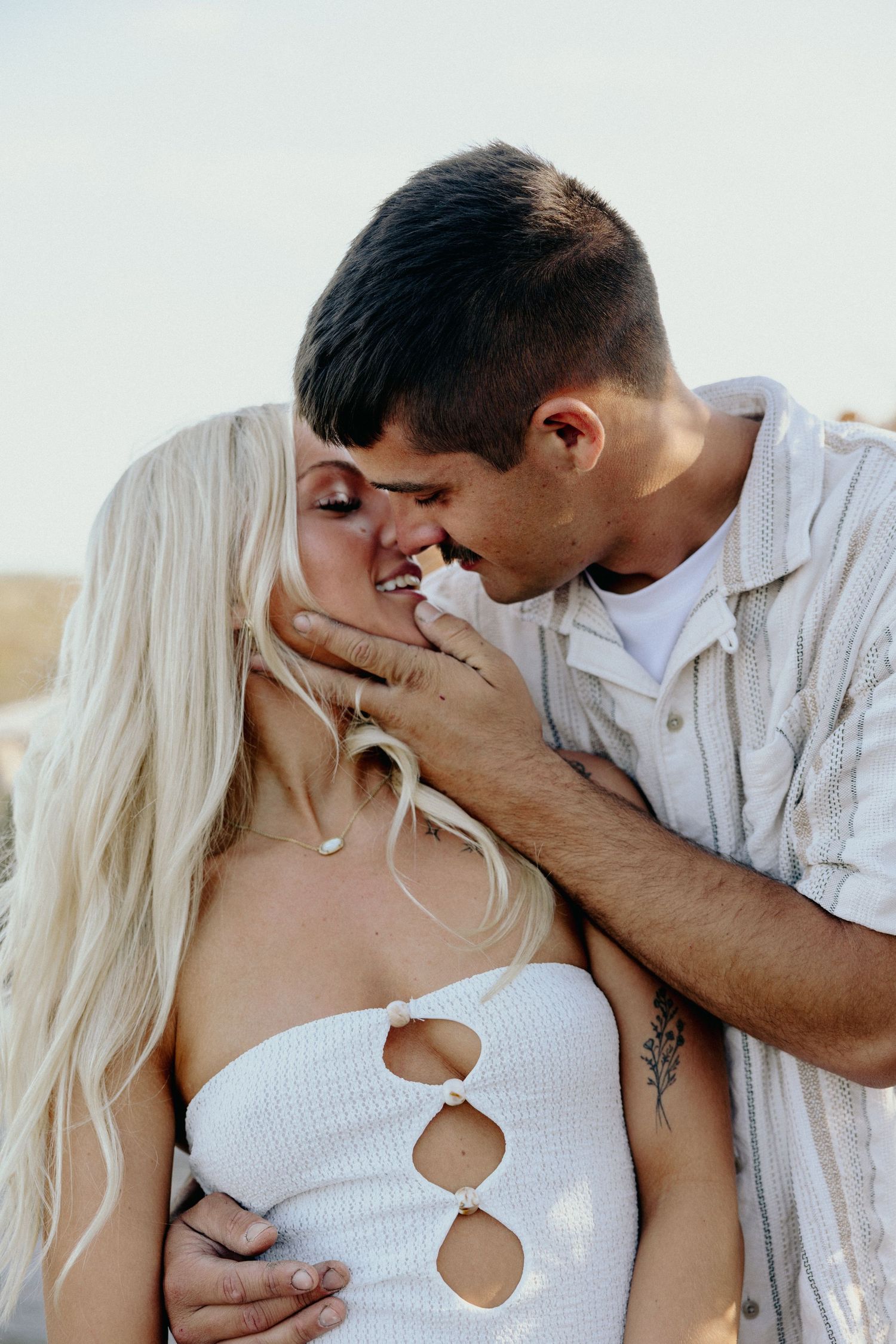 A romantic couple shares an intimate moment, the man tenderly cupping the smiling blonde woman's face as they lean in to kiss.