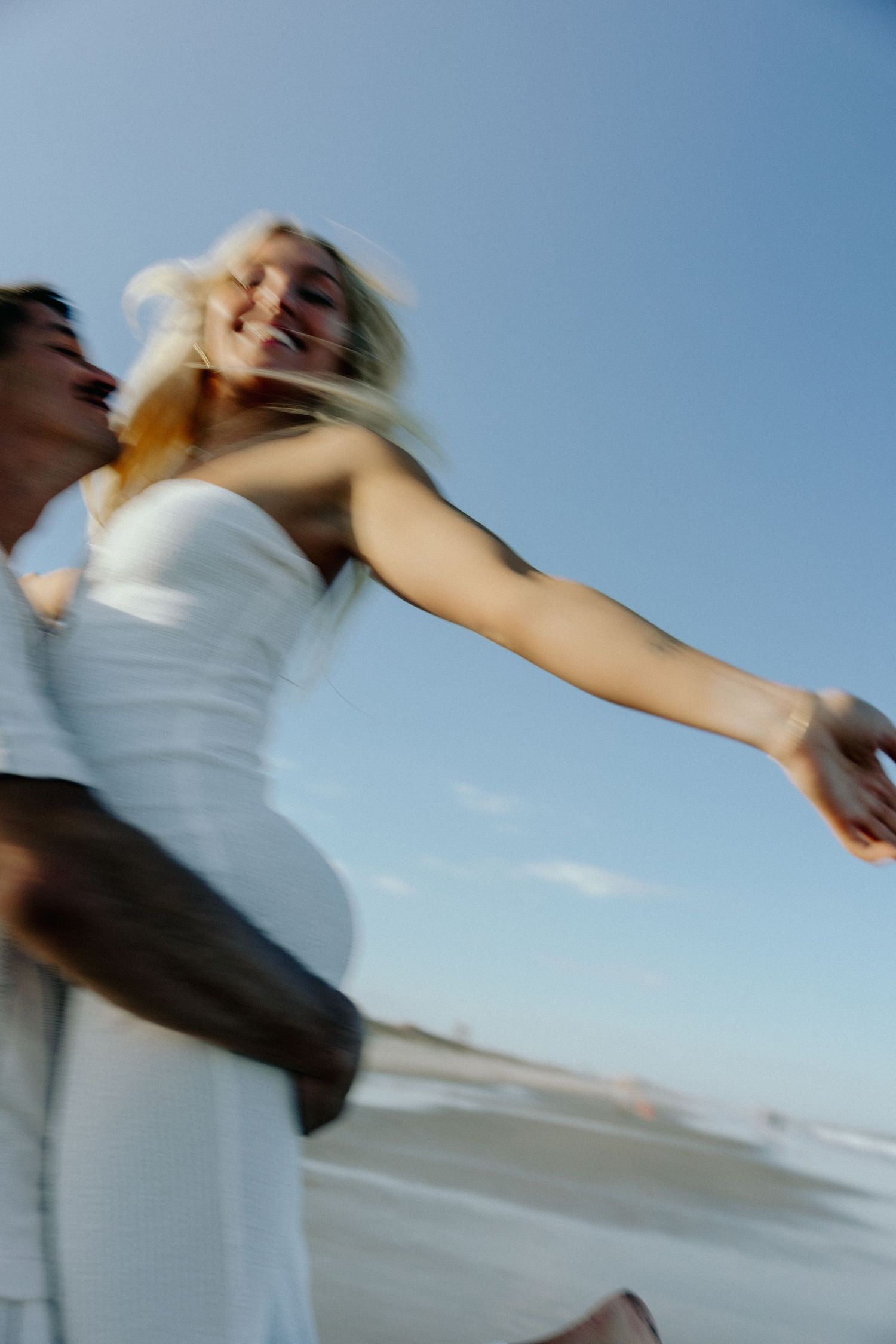 A joyful couple in white attire spinning on a beach, with the woman laughing and arms outstretched under a clear blue sky.