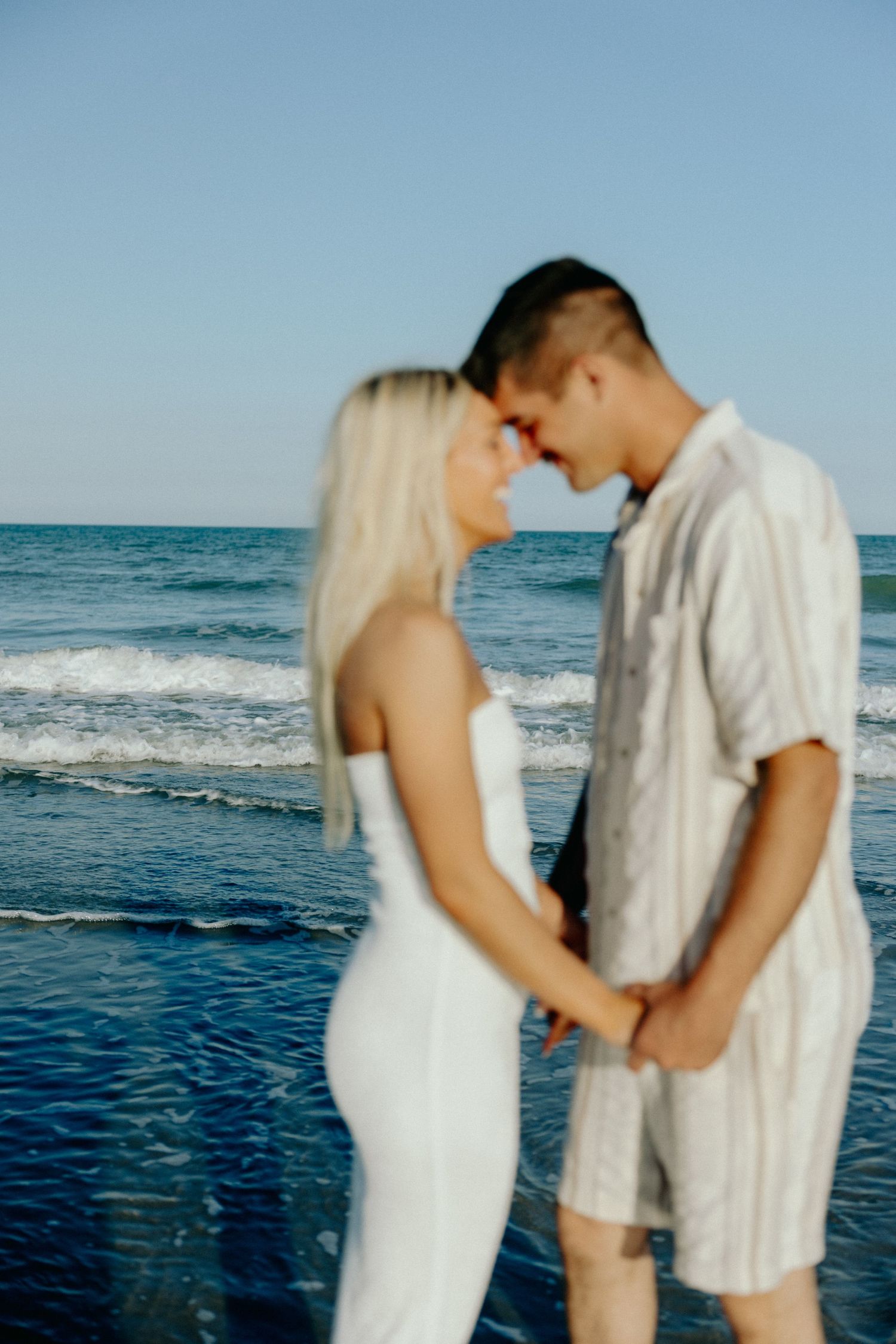 Romantic couple holding hands and touching foreheads on a beach with blue ocean waves in the background.