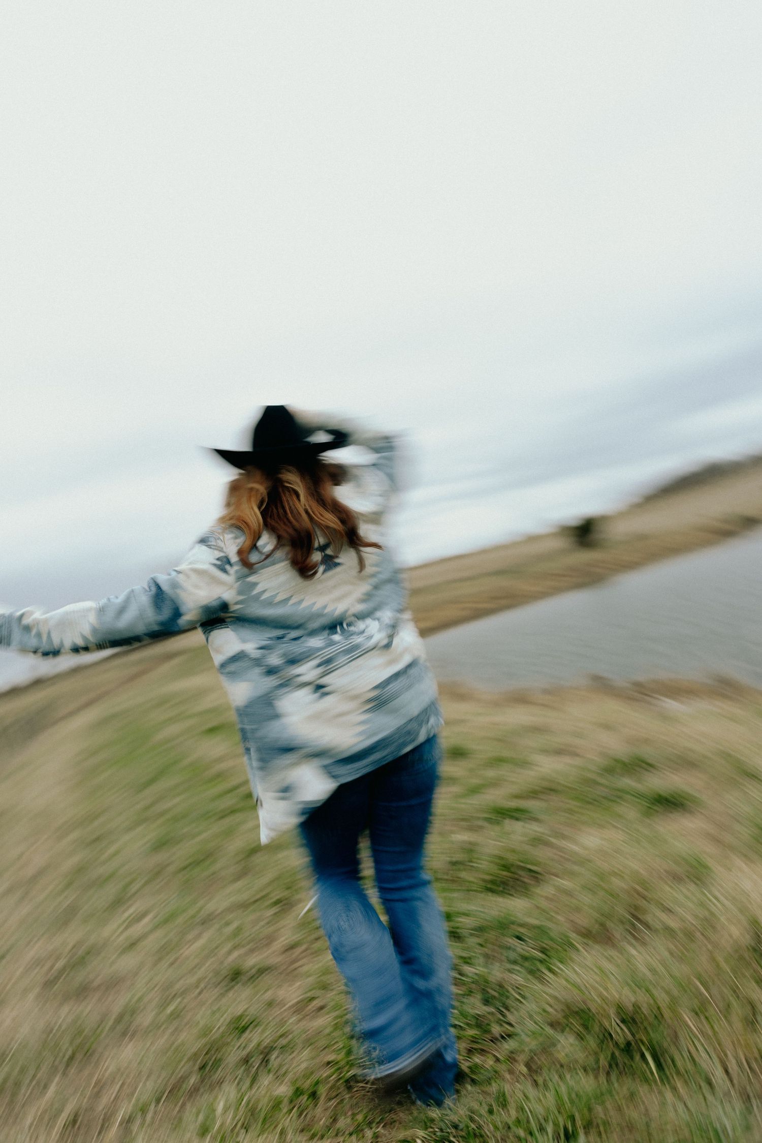 A woman in a cowboy hat, patterned jacket, and jeans spins outdoors near a lake on an overcast day.
