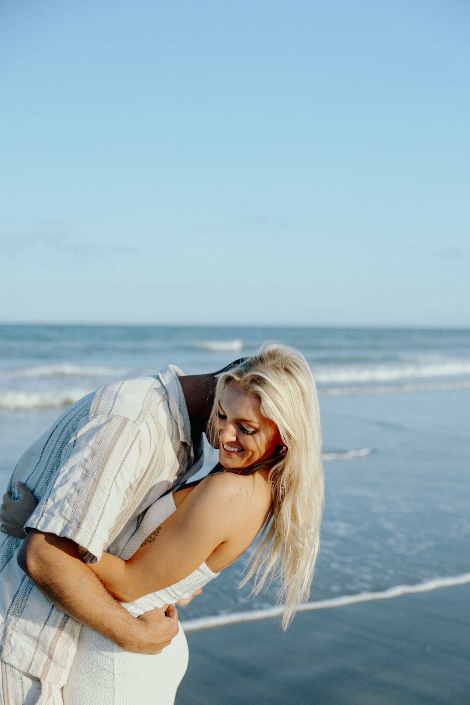 A smiling blonde woman embraces her partner on a sunny beach with ocean waves in the background.