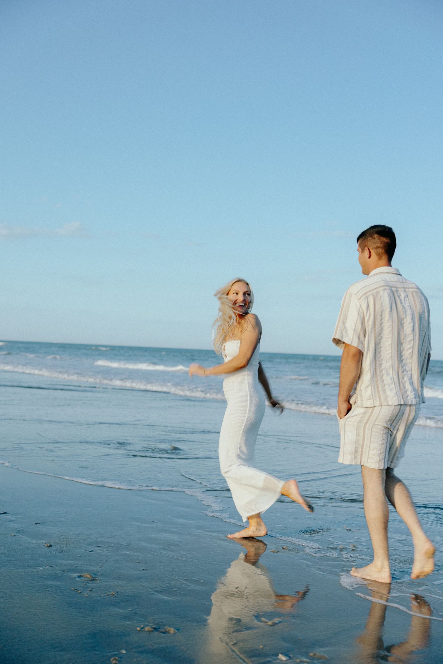 A joyful couple walks barefoot on a wet sandy beach, woman in white dress laughing, waves in background.