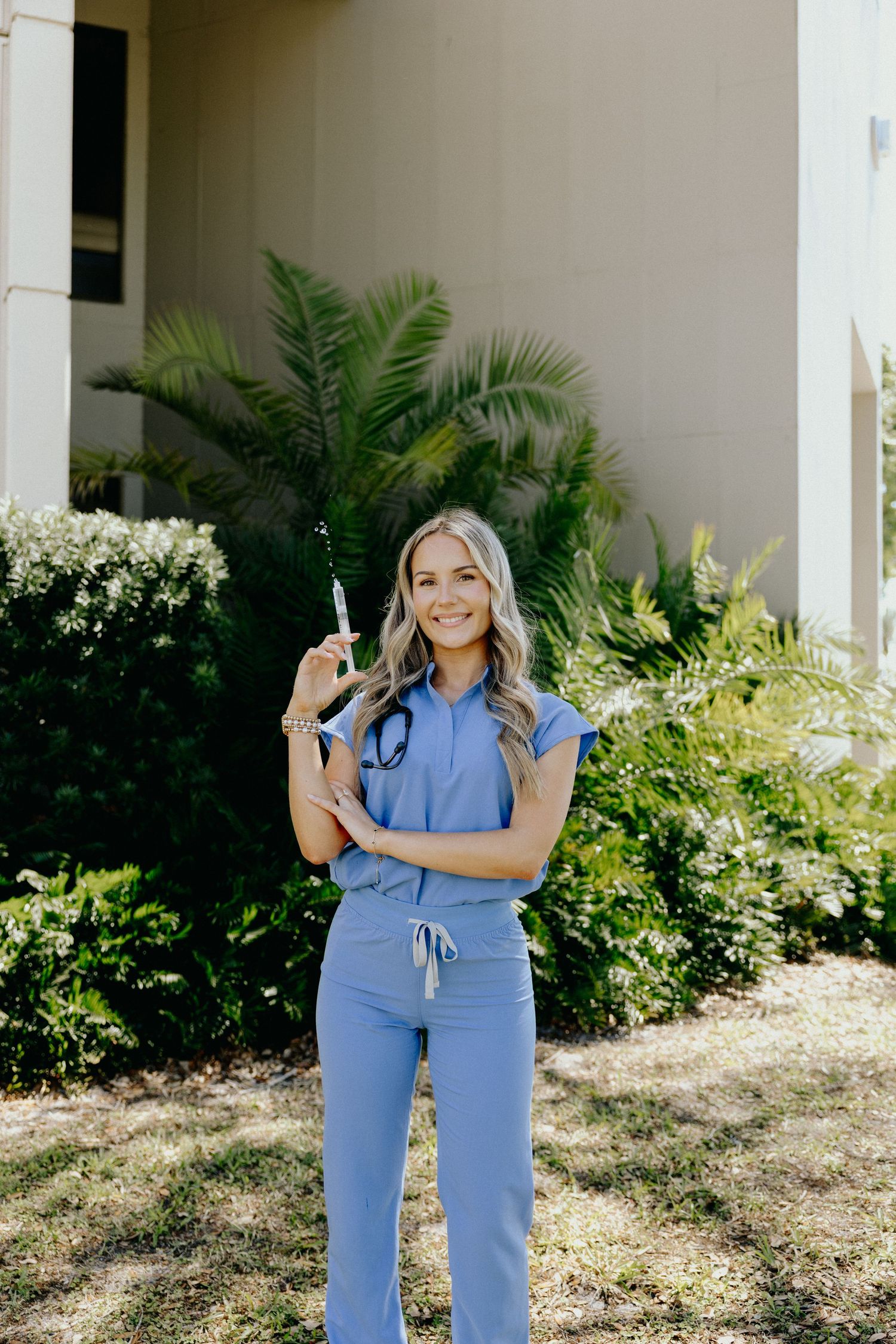 Smiling female nurse in blue scrubs holding a syringe outdoors with tropical plants in background.