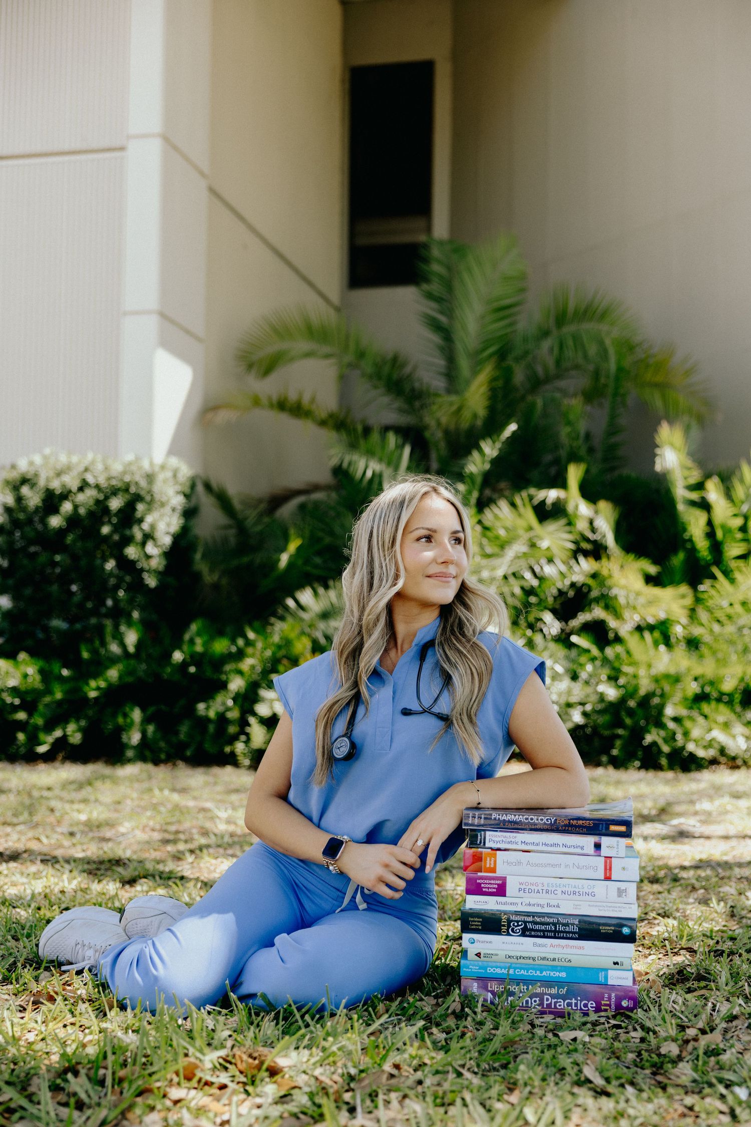 Nursing student in blue scrubs sitting on grass with stack of medical textbooks outdoors.