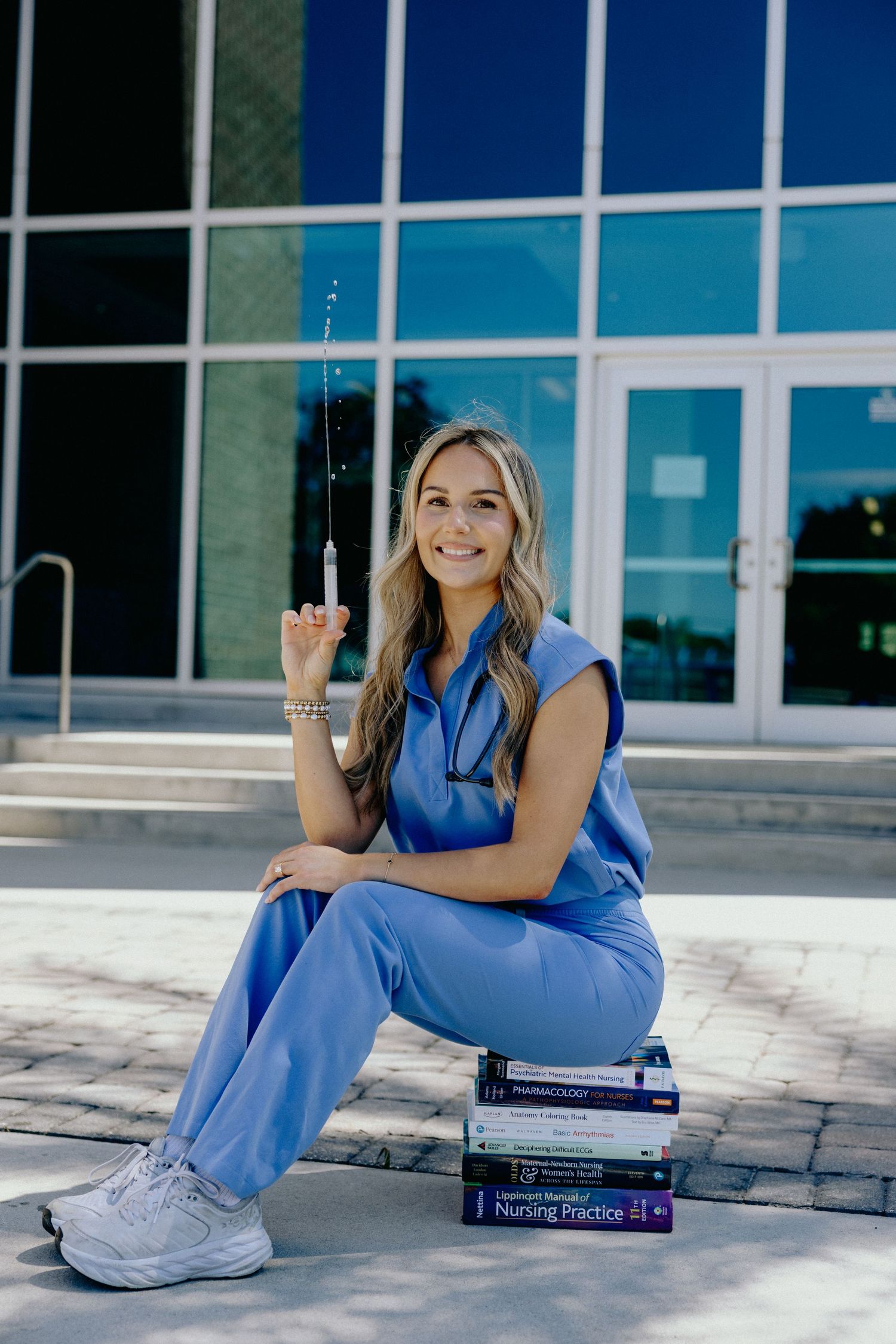 Happy nursing student in blue scrubs holding syringe while sitting on medical textbooks outside.
