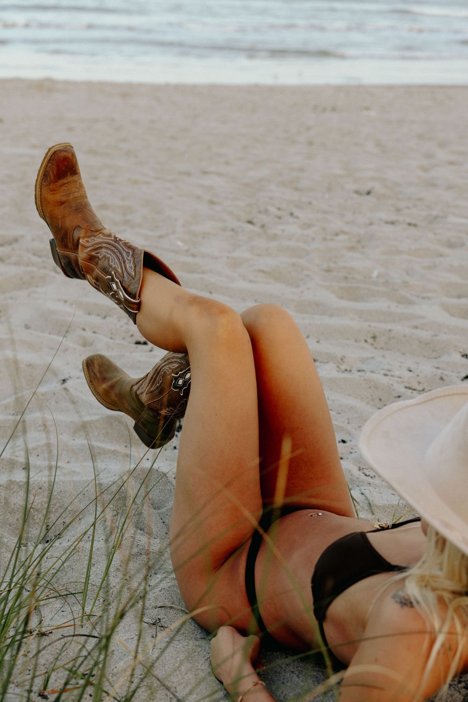 Woman in bikini and cowboy boots relaxing on sandy beach with ocean in background.