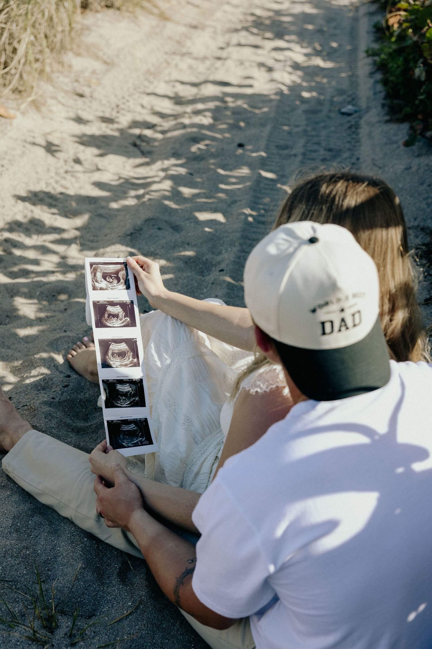 Couple sitting on sandy beach reviewing ultrasound photos, man wearing white DAD cap, pregnancy announcement moment.