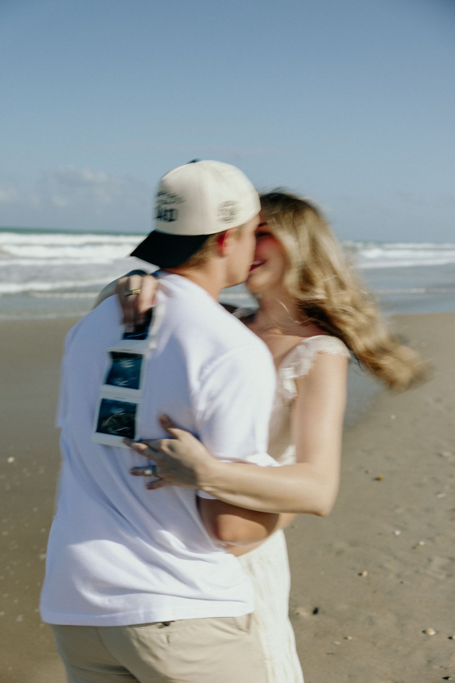 Couple kissing on beach holding ultrasound photos, pregnancy announcement, ocean waves in background, sunny day.