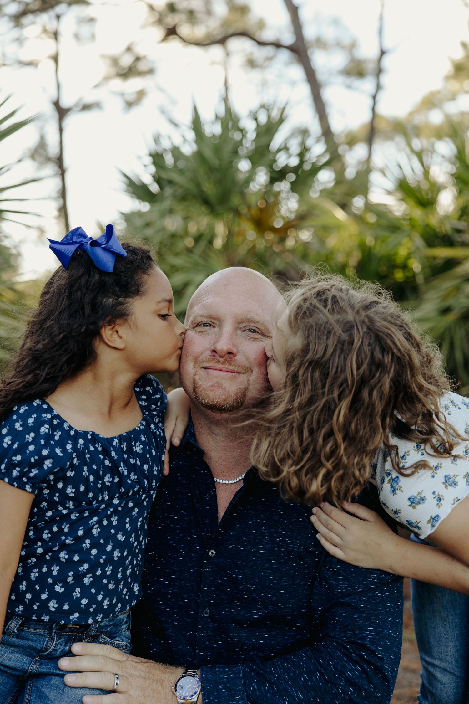 A dad smiles as two young girls kiss him on both cheeks outdoors.