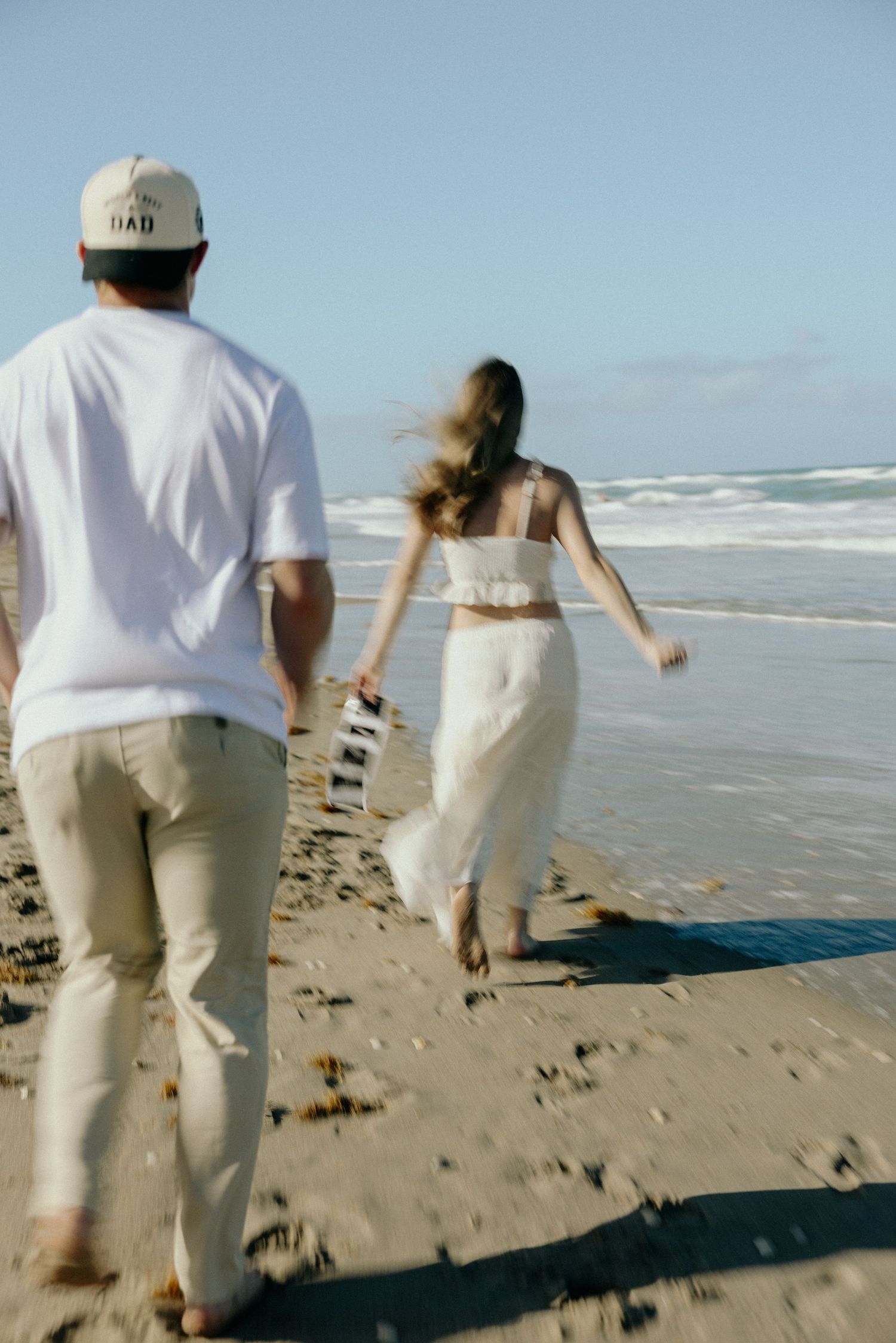 A couple runs barefoot along a sunny beach shoreline, the man in white tee and cap, woman in flowing white dress.