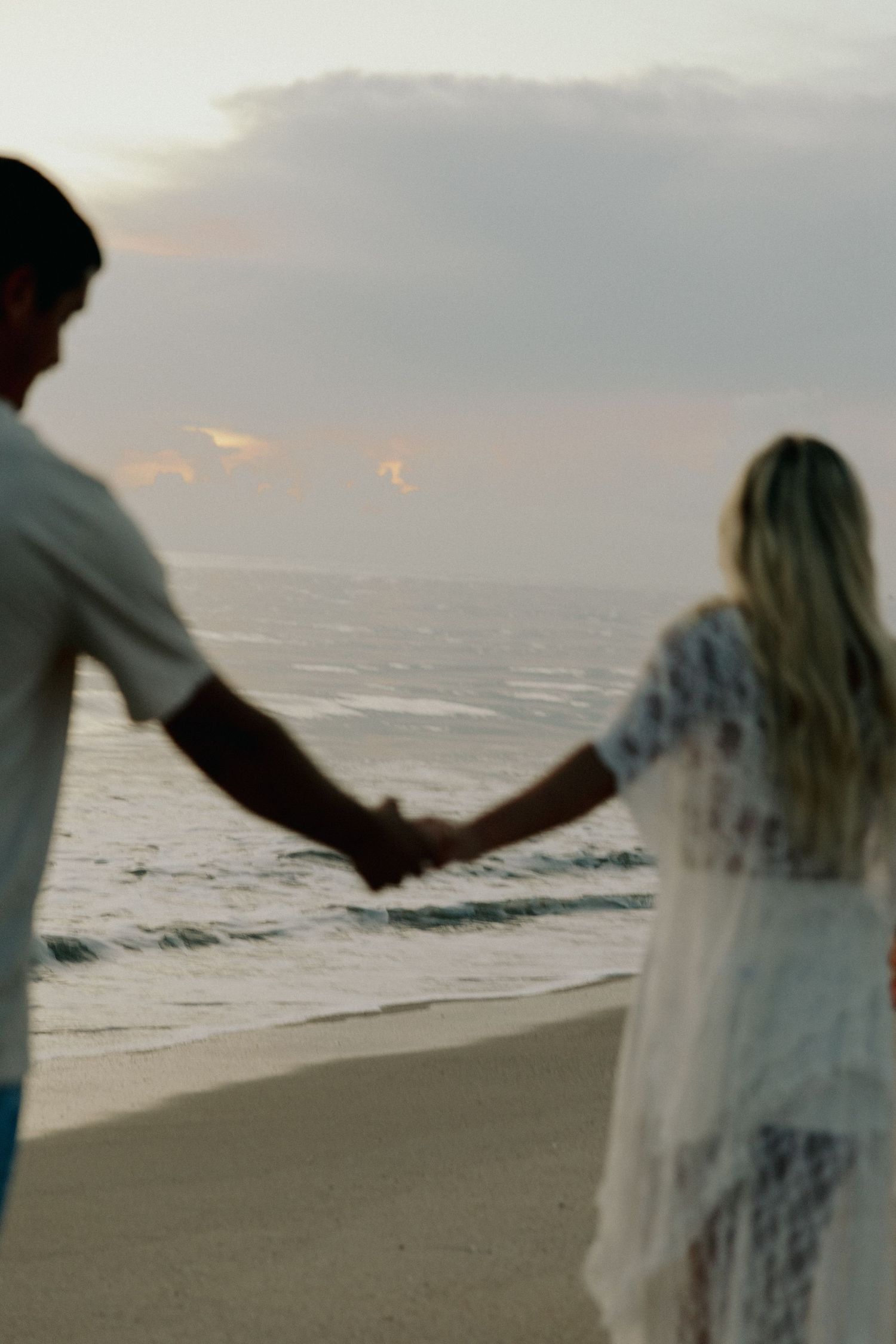 Couple holding hands at beach during sunset with colorful cloudy sky.