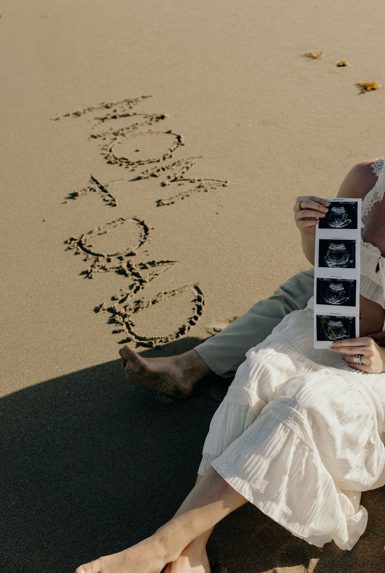 Pregnant woman in white dress sits on sandy beach holding ultrasound photos beside writing in the sand.