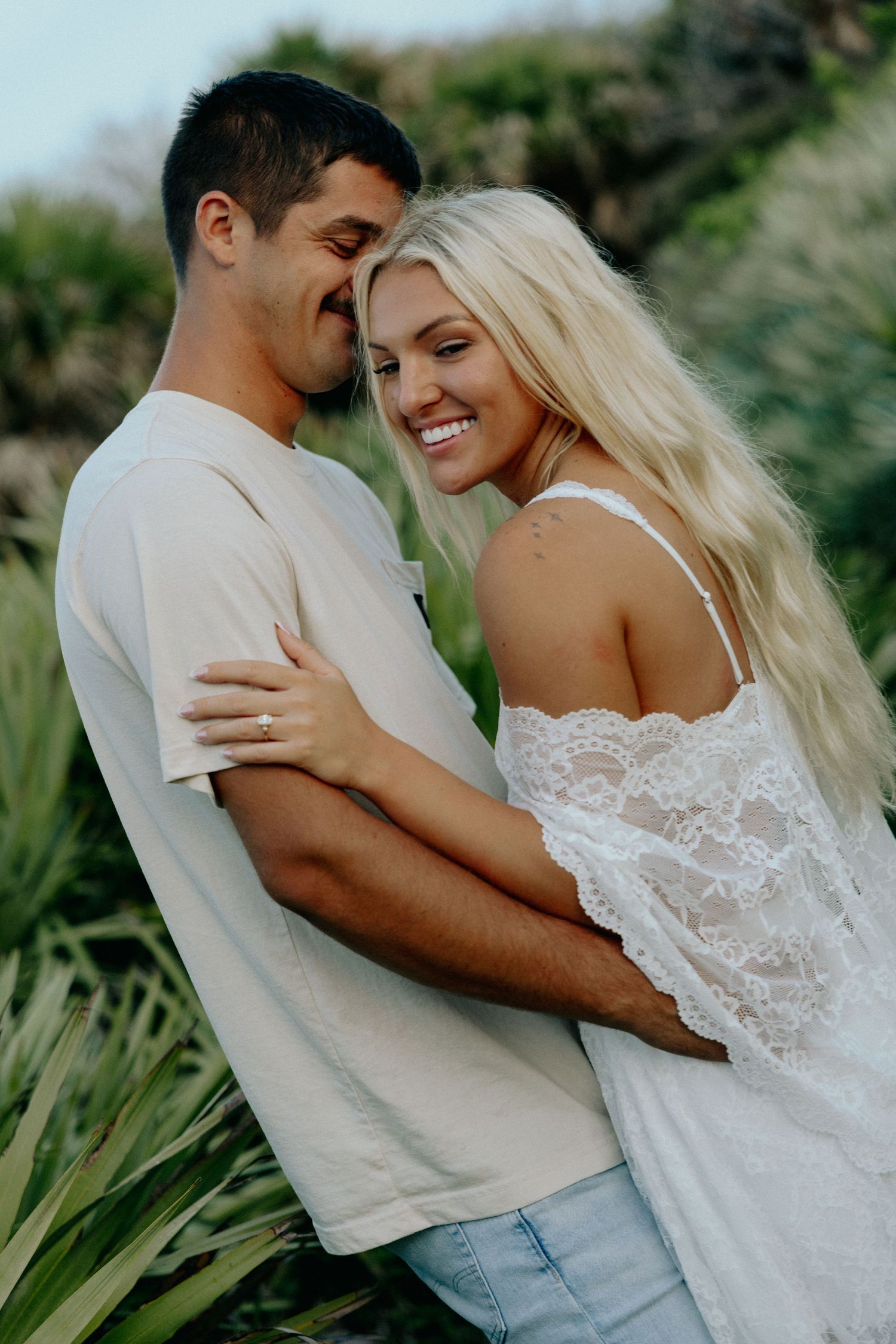 Smiling blonde woman embracing man in beige shirt surrounded by tropical greenery.