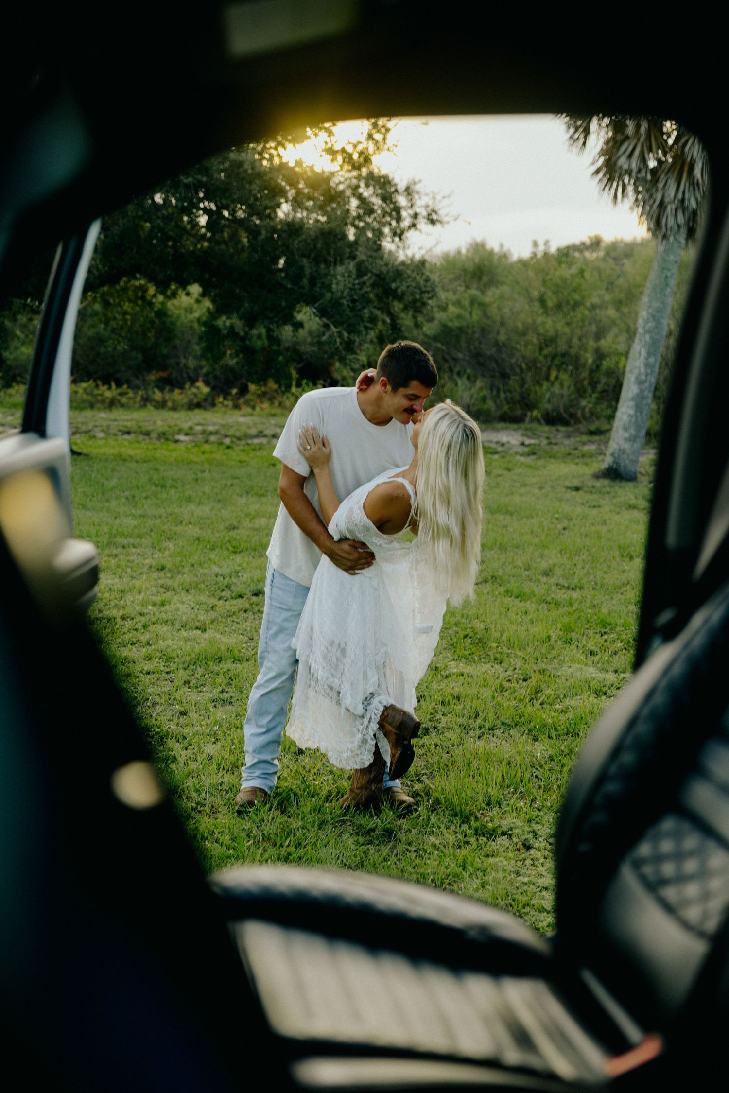Couple kissing in sunlit green field photographed through open car trunk.