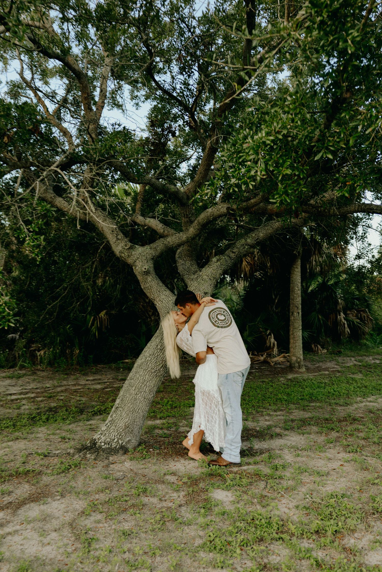 Couple sharing romantic kiss leaning against tree in lush outdoor setting.