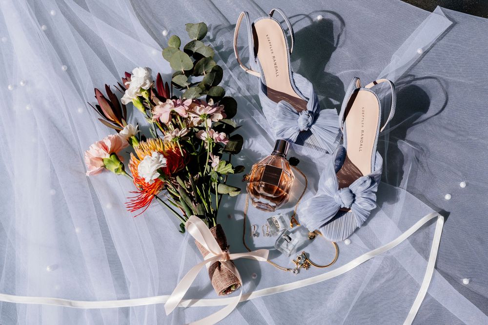 Elegant wedding accessories including perfume bottle, pink heels, and floral bouquet arranged on light fabric.