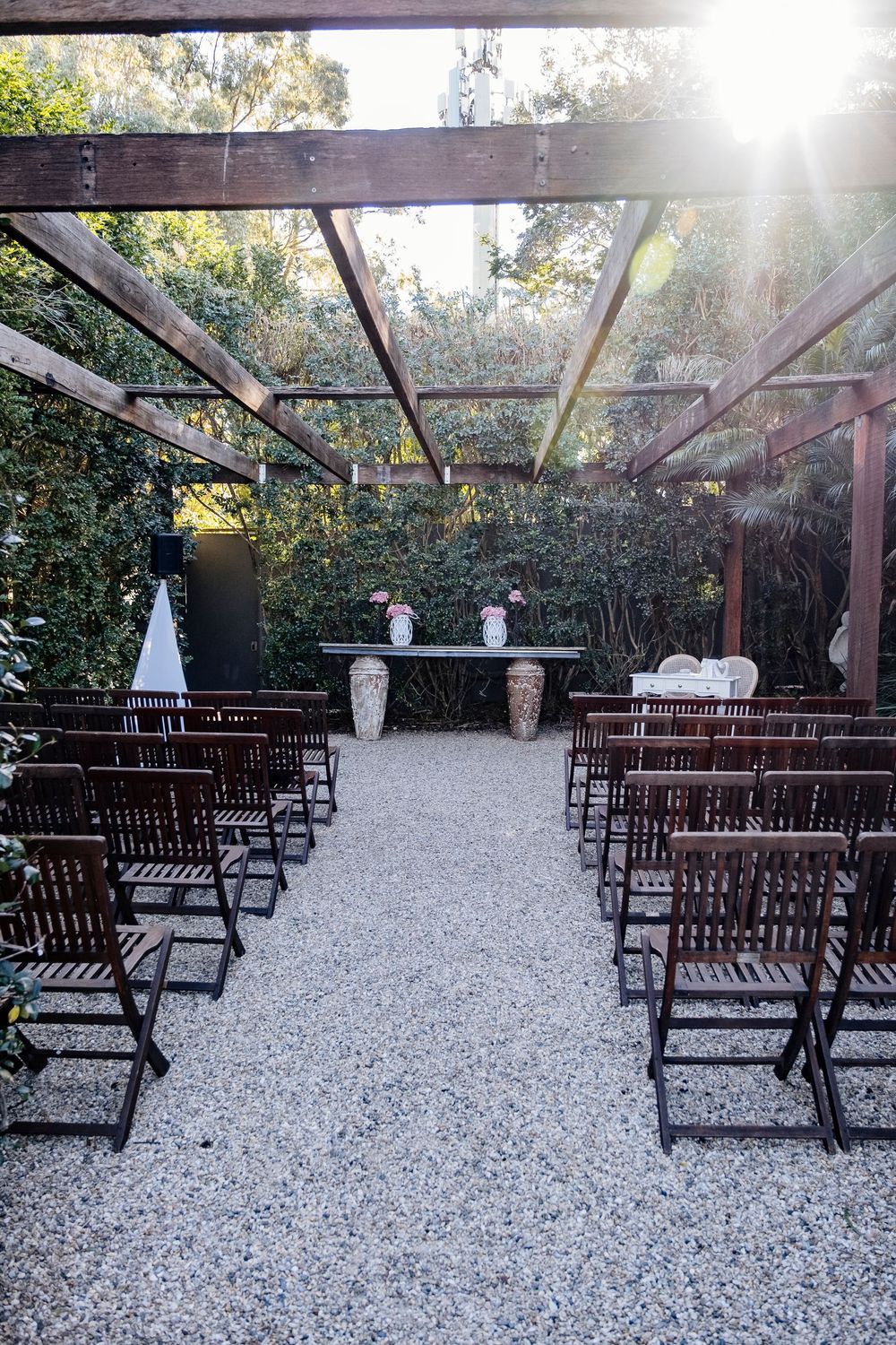 Rustic outdoor wedding ceremony setup with wooden benches and white decorative urns on gravel under a natural canopy.
