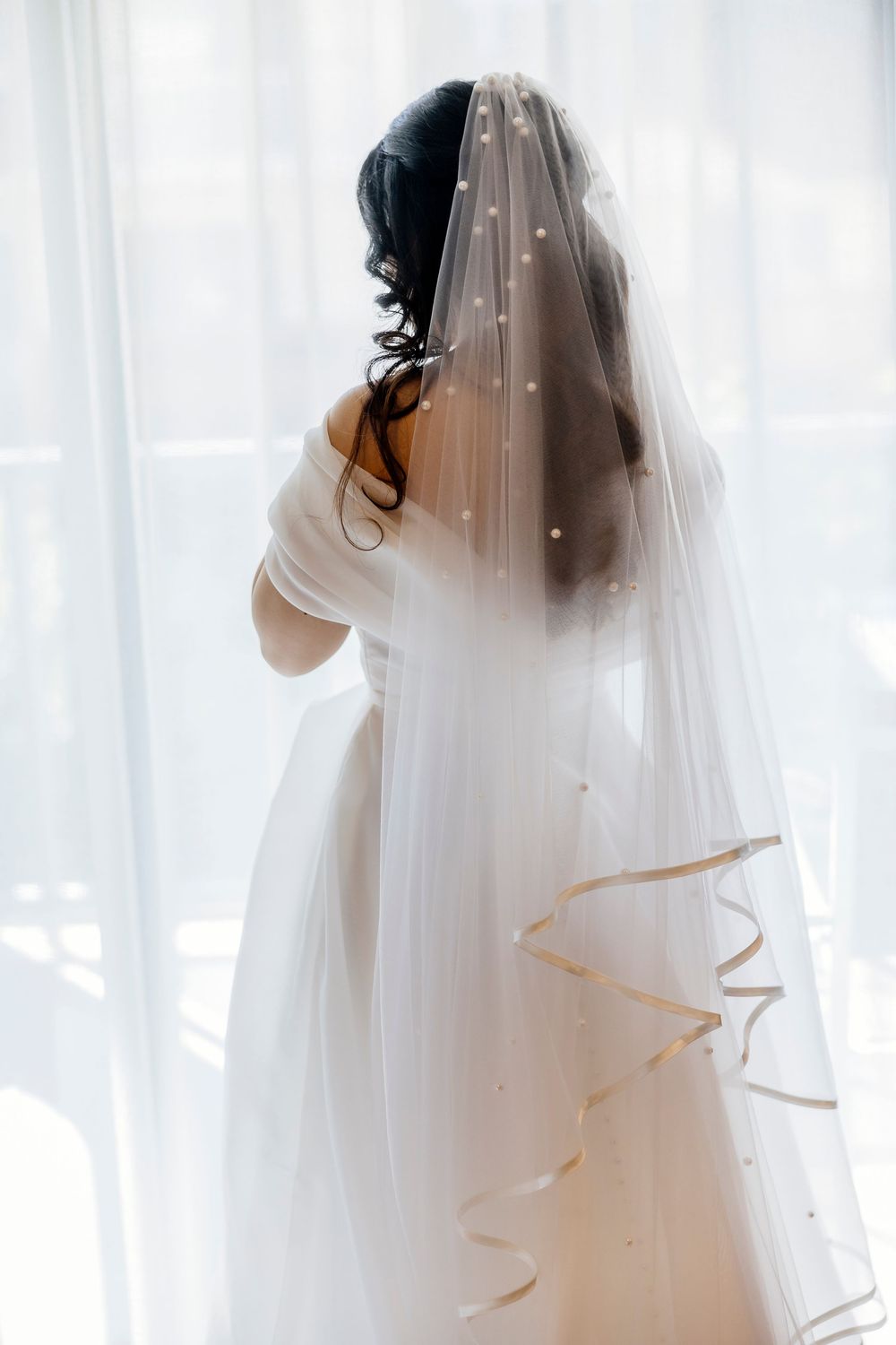A flowing white wedding veil with delicate trim cascades down against sheer curtains in natural light.