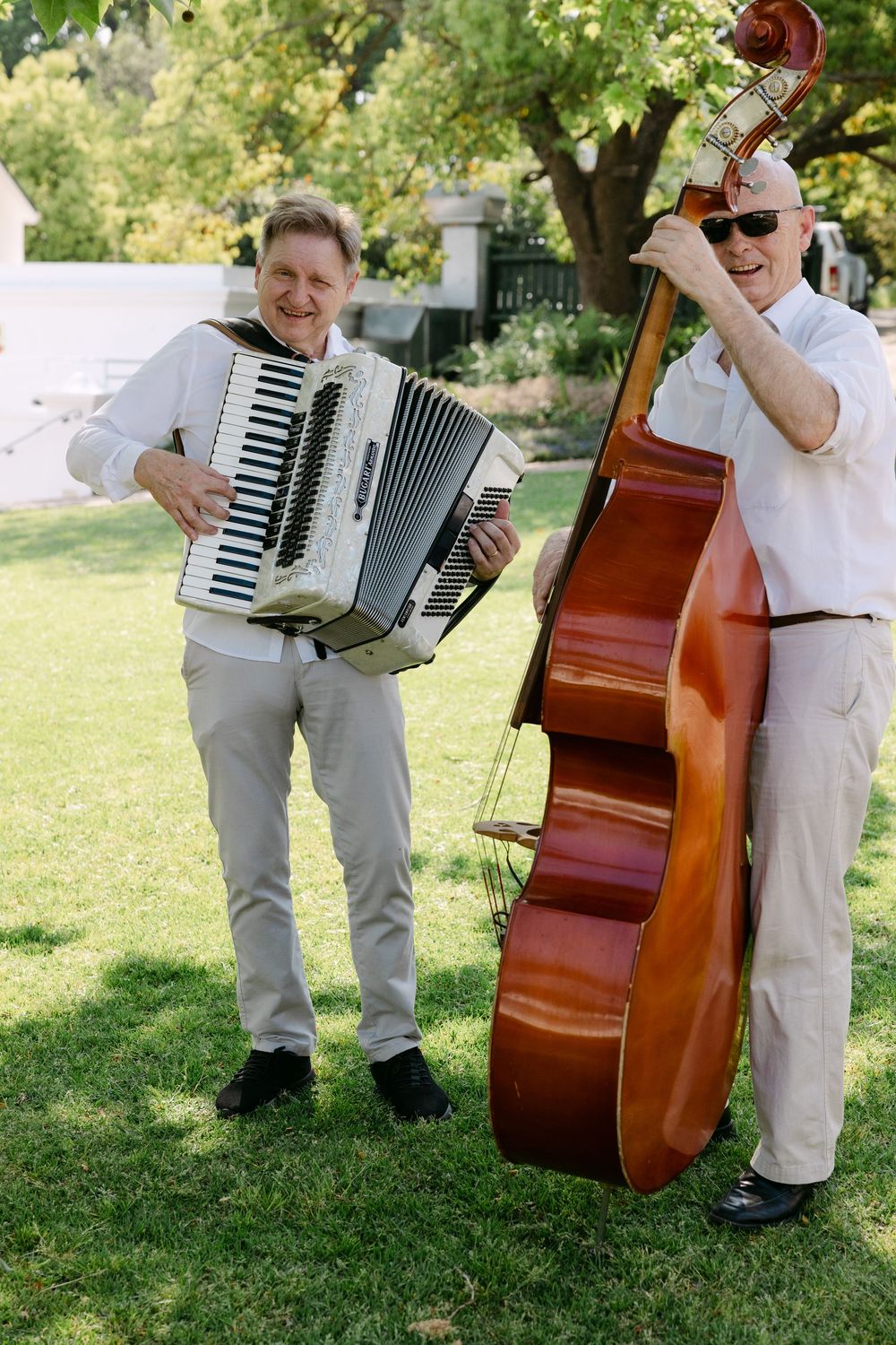 Musicians perform outdoors with an accordion and upright bass on a sunny lawn.