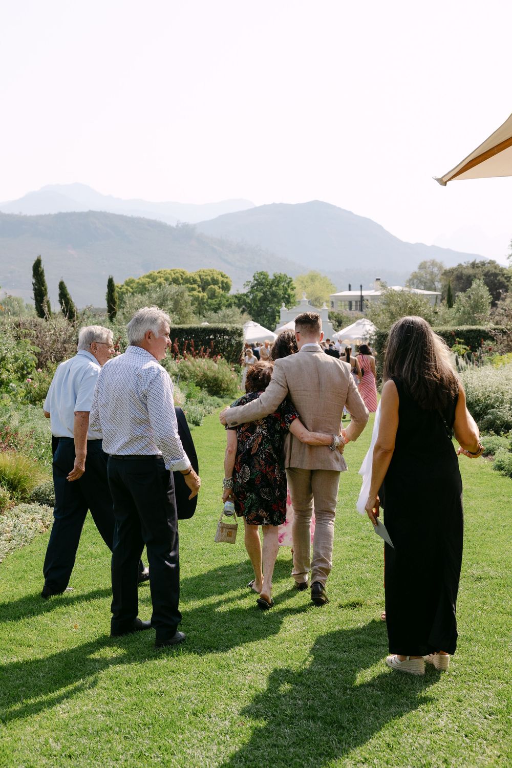 Wedding guests gather on a lush green lawn with mountains in the background during an outdoor celebration.