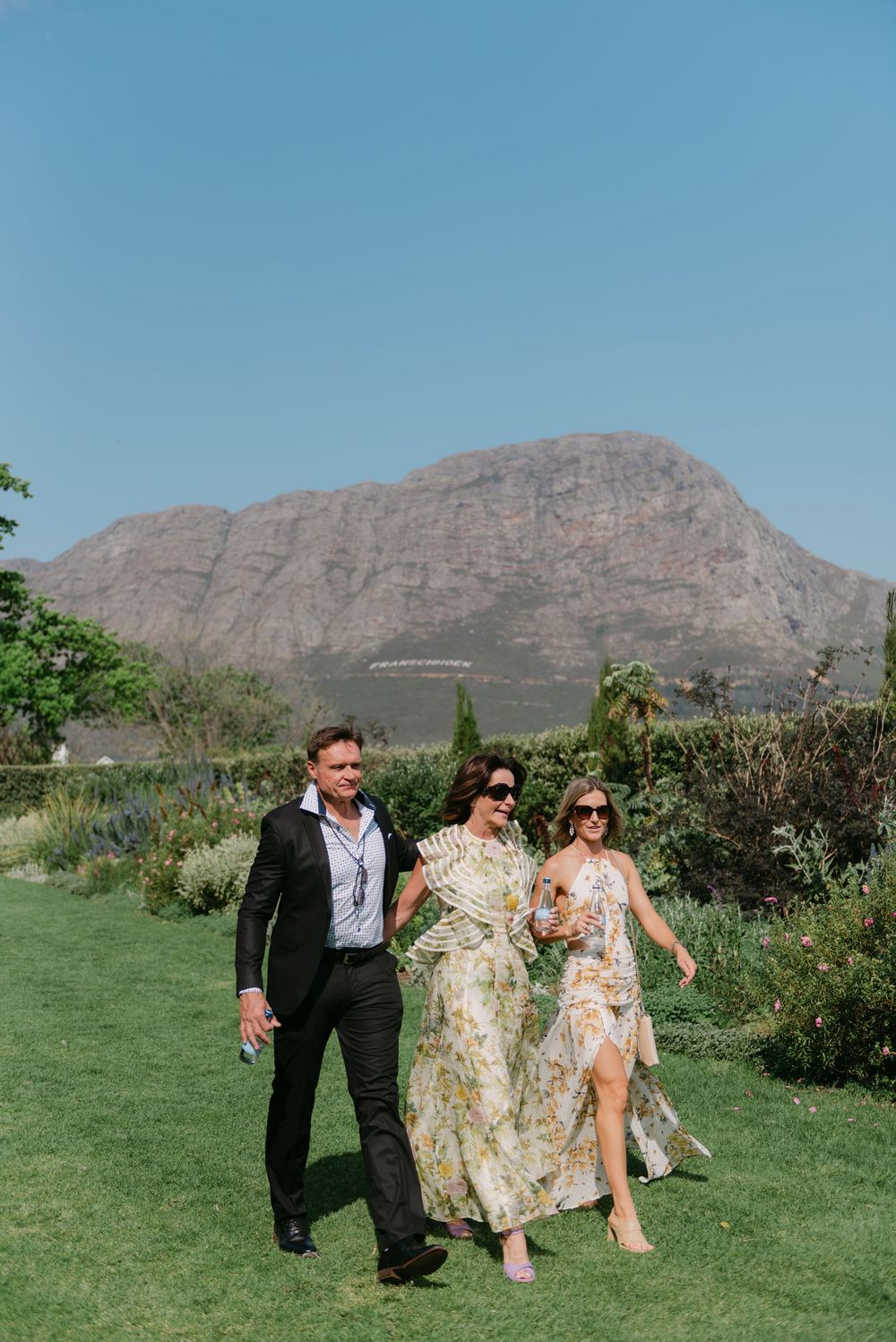 Wedding party members walk and laugh together on a grassy lawn with mountains in the background.