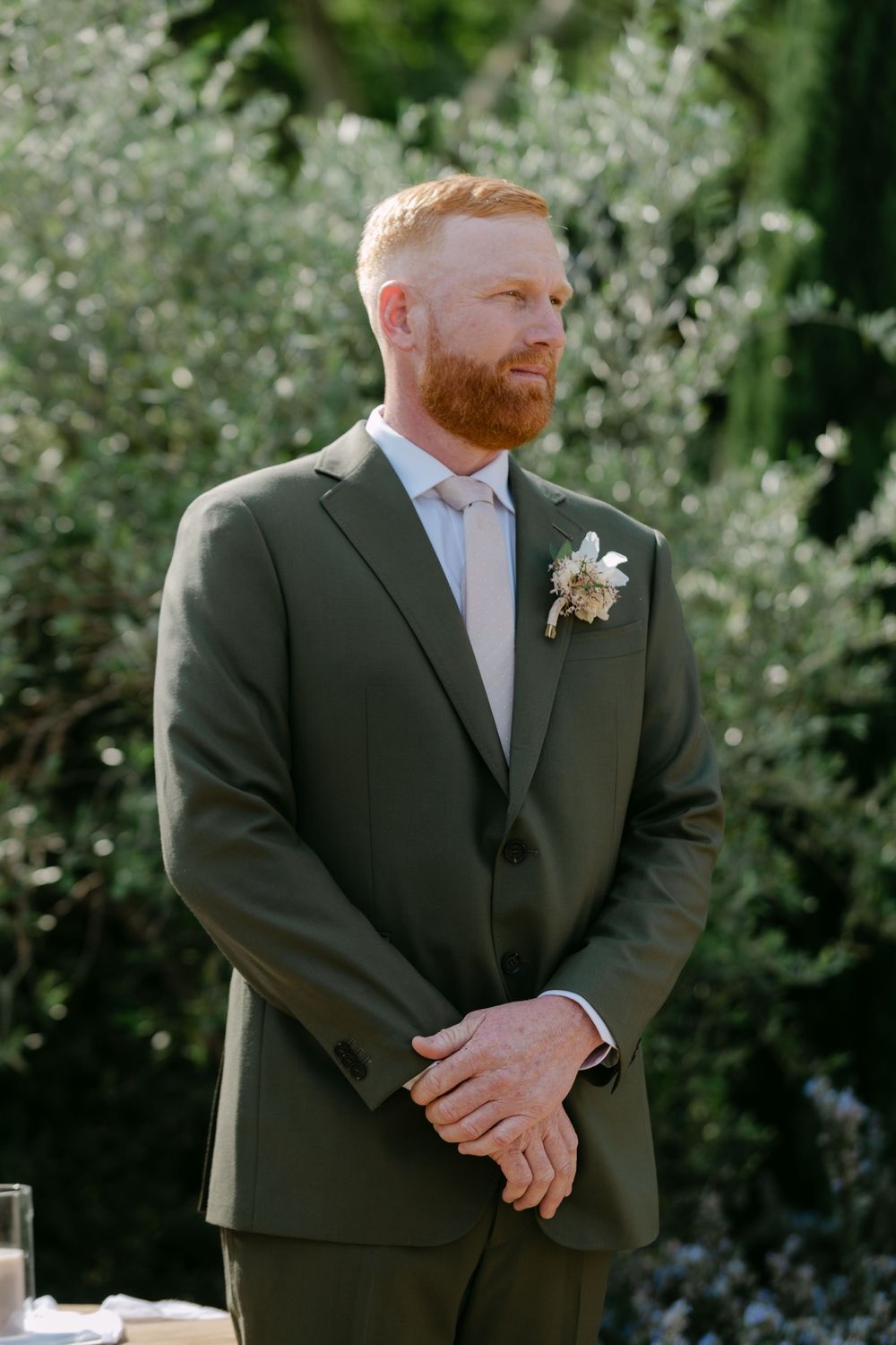 A groom in a dark suit stands in a garden setting with white flowering trees in the background.