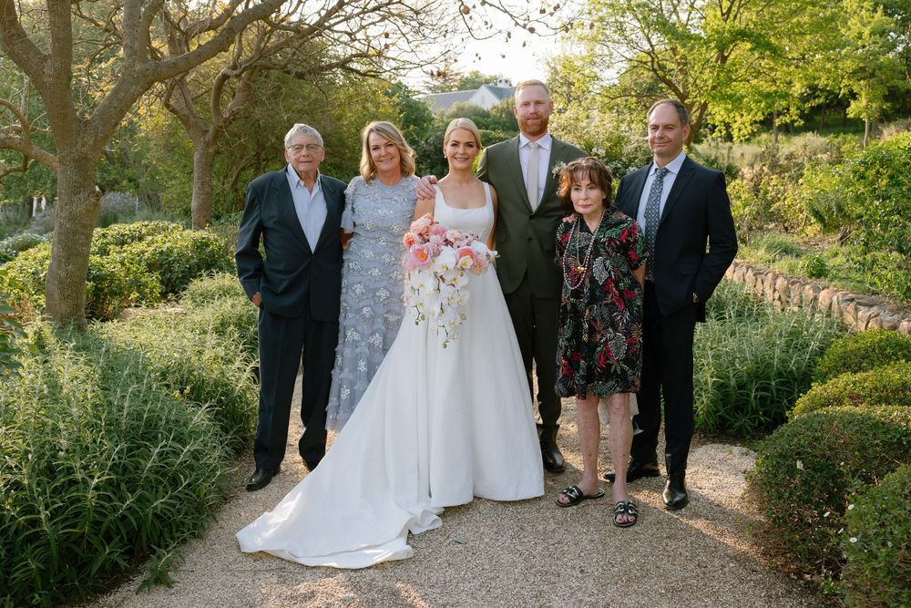 A scenic outdoor garden wedding ceremony with guests gathered among lush greenery and flowering plants in natural light.