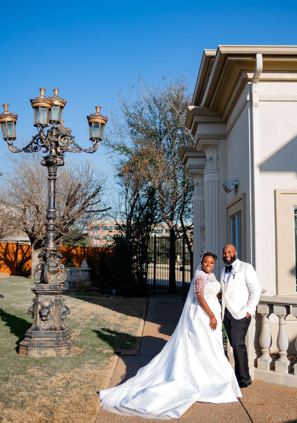 A couple in wedding attire poses for romantic outdoor photos near ornate stone pillars and lampposts.