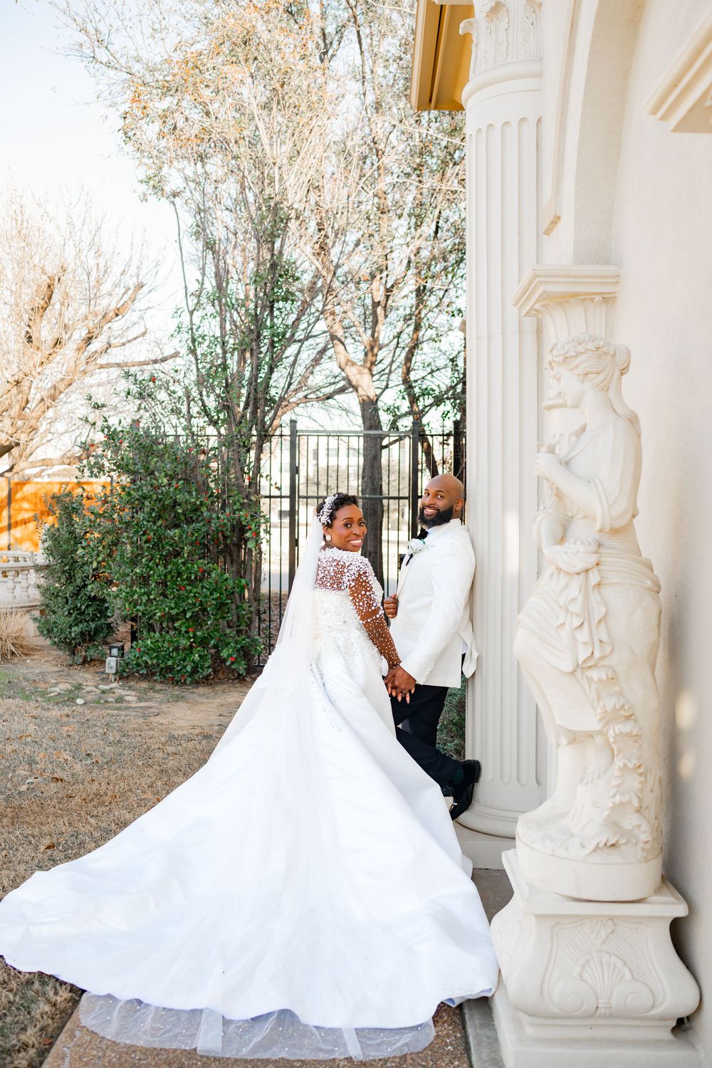 A couple in wedding attire poses elegantly by ornate white columns and greenery on their special day.