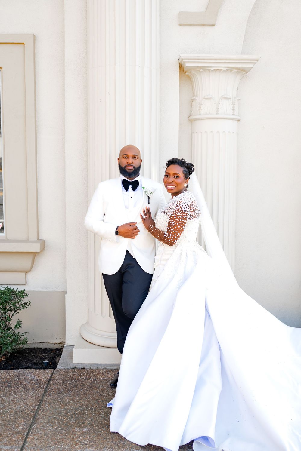 A couple poses together on their wedding day, with the bride in a stunning white gown and the groom in a white tuxedo jacket.
