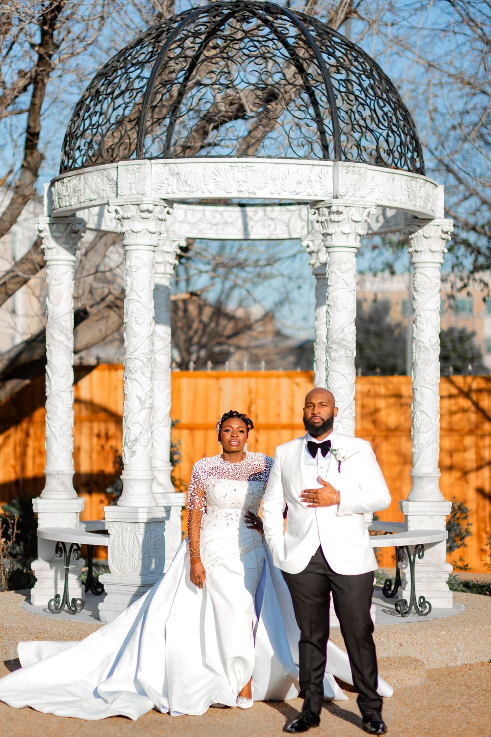 A romantic wedding photoshoot with white columns and orange fence backdrop in an outdoor setting.