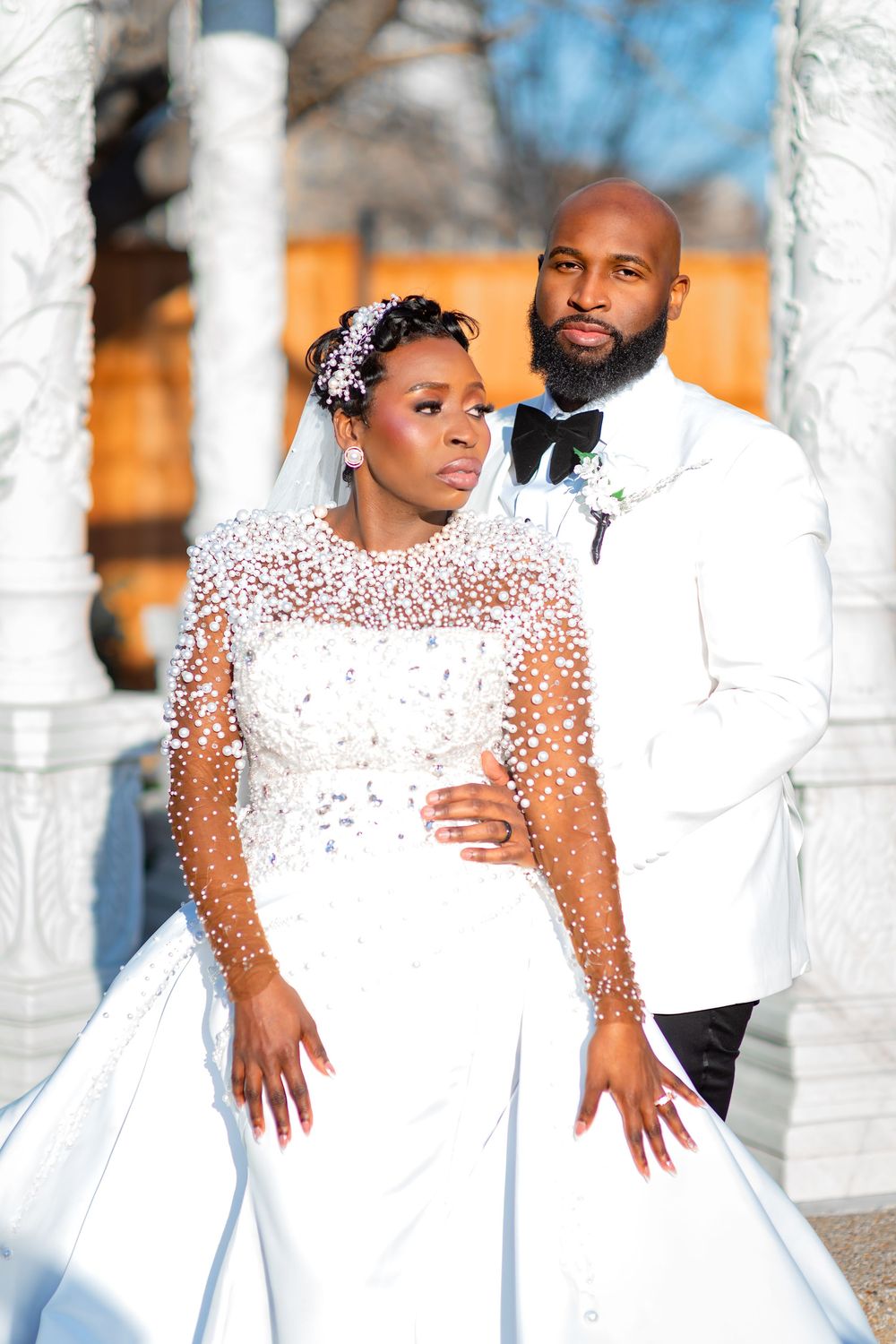 A couple in matching white outfits poses together in a romantic photoshoot against white columns outdoors.