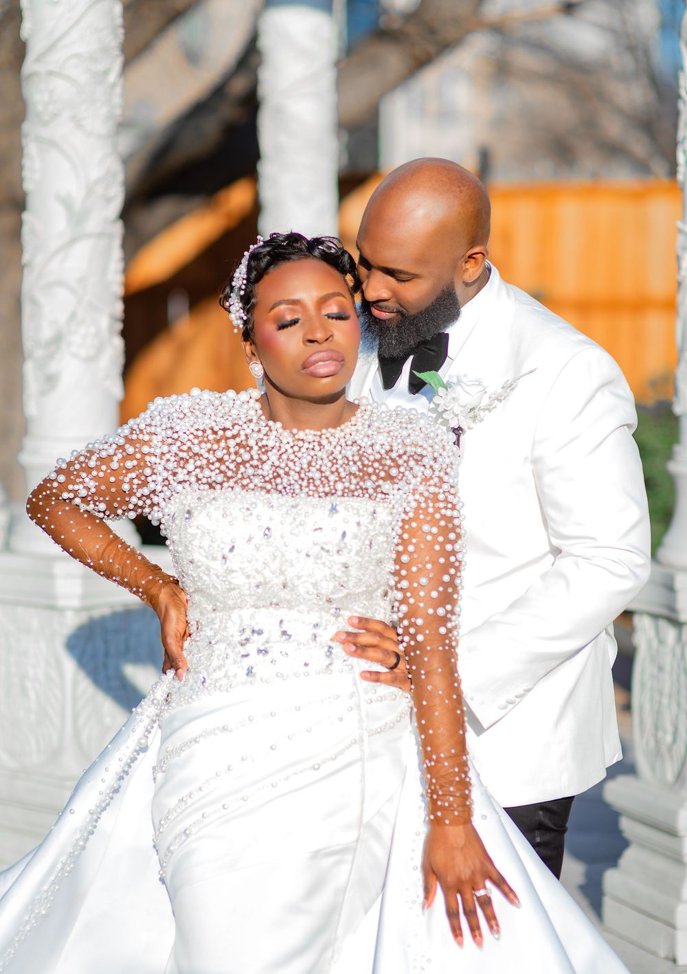 A couple in elegant white attire poses together in a romantic outdoor photoshoot with white pillars in the background.