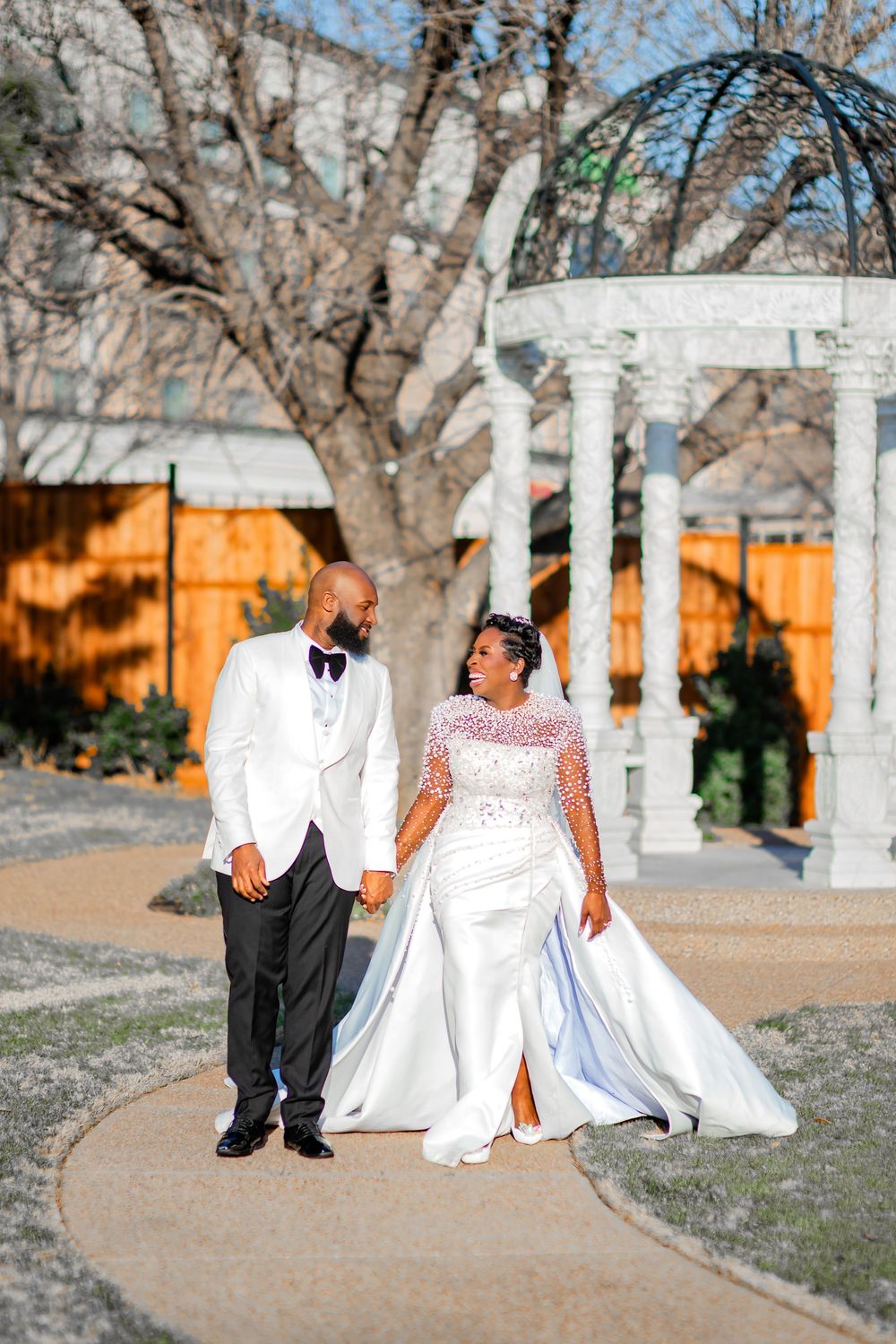 A wedding couple poses together on a path with white pillars and bare trees in the background.