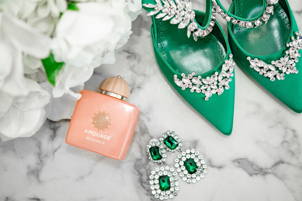 Green and white wedding accessories including emerald jewelry, satin ribbons, and floral arrangements on a marble surface.