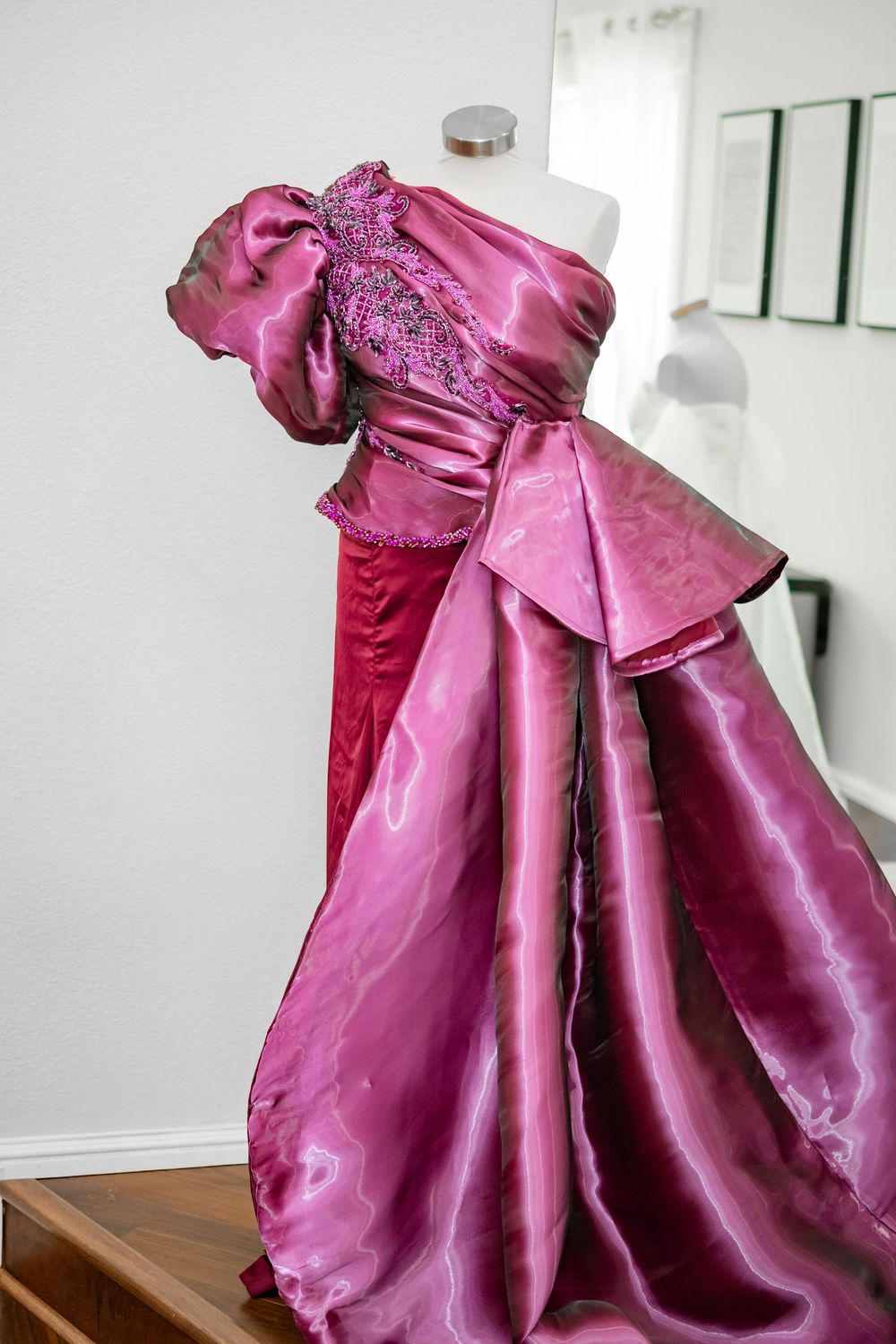 Elegant magenta satin formal dress with dramatic bow detail and flowing skirt displayed against white wall.