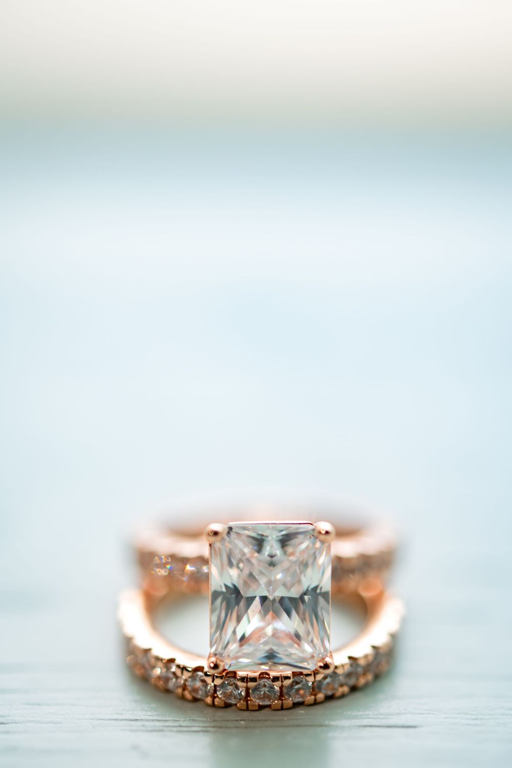 Rose gold engagement ring with princess cut diamond and delicate band sparkles against light blue background.