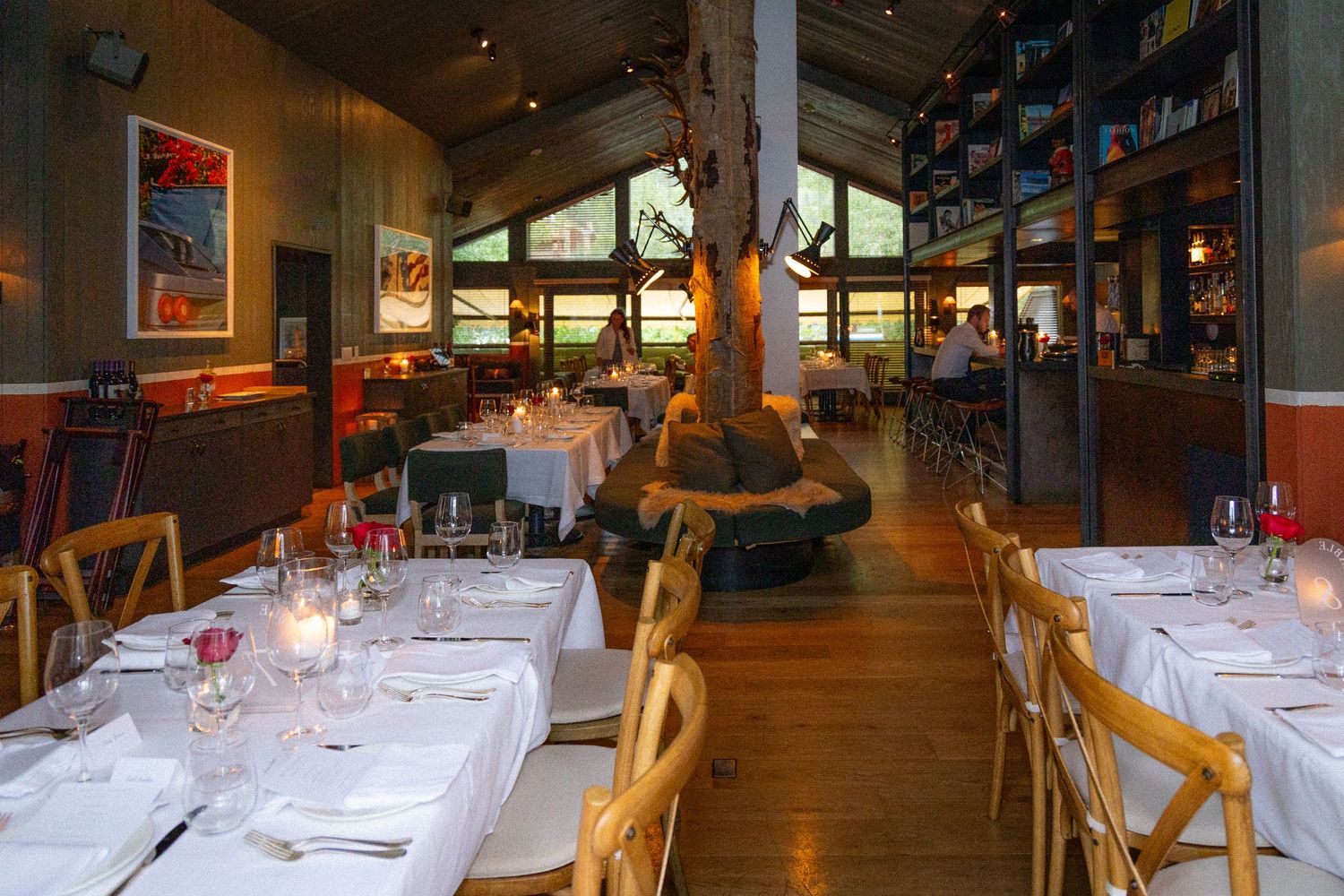 Long wooden dining tables with white tablecloths set for service in a cozy restaurant interior with warm lighting.