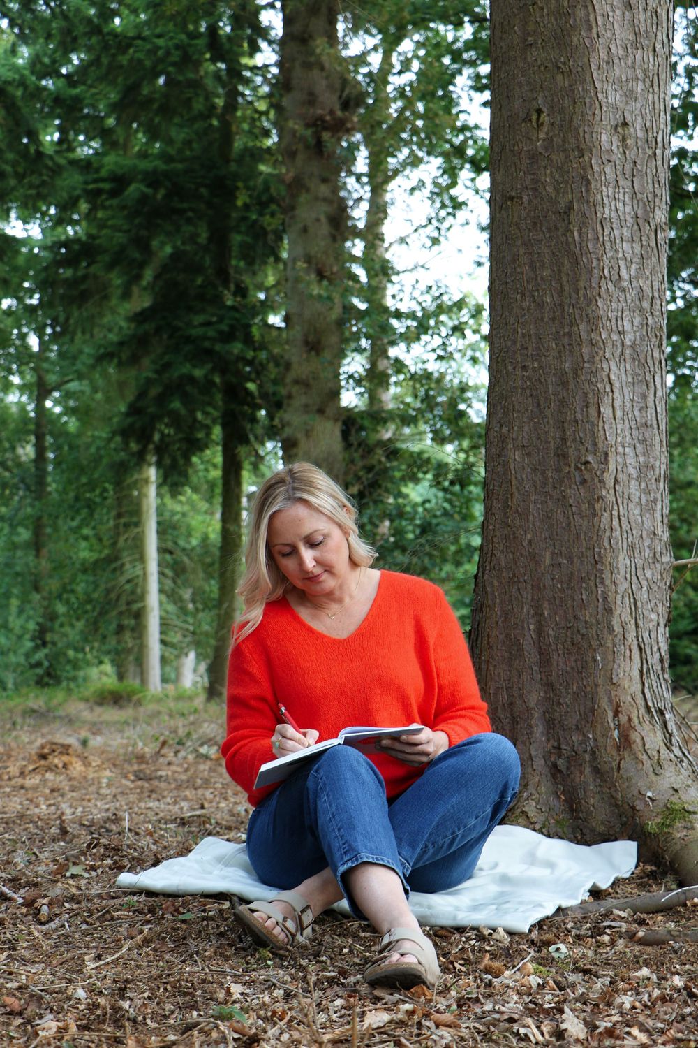 Person in bright orange sweater and jeans sits on white blanket under tall trees writing in notebook.